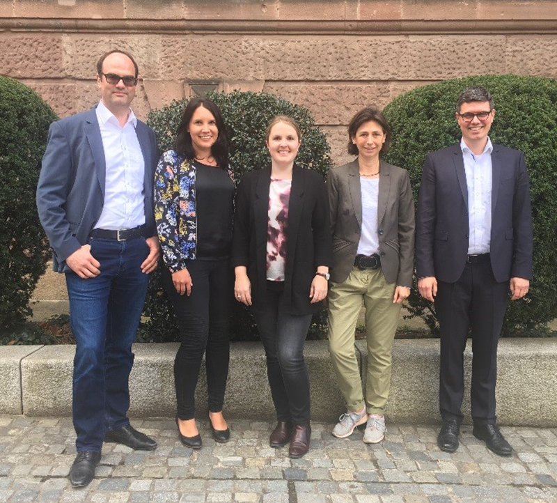 Fünf Menschen stehen nebeneinander im Freien, hinter ihnen eine Steinmauer und Büsche. Sie sind halbformell gekleidet und lächeln in die Kamera. Der Hintergrund vermittelt ein lebendiges Campus-Gefühl und erinnert an den lebendigen Geist der Hochschule Coburg.