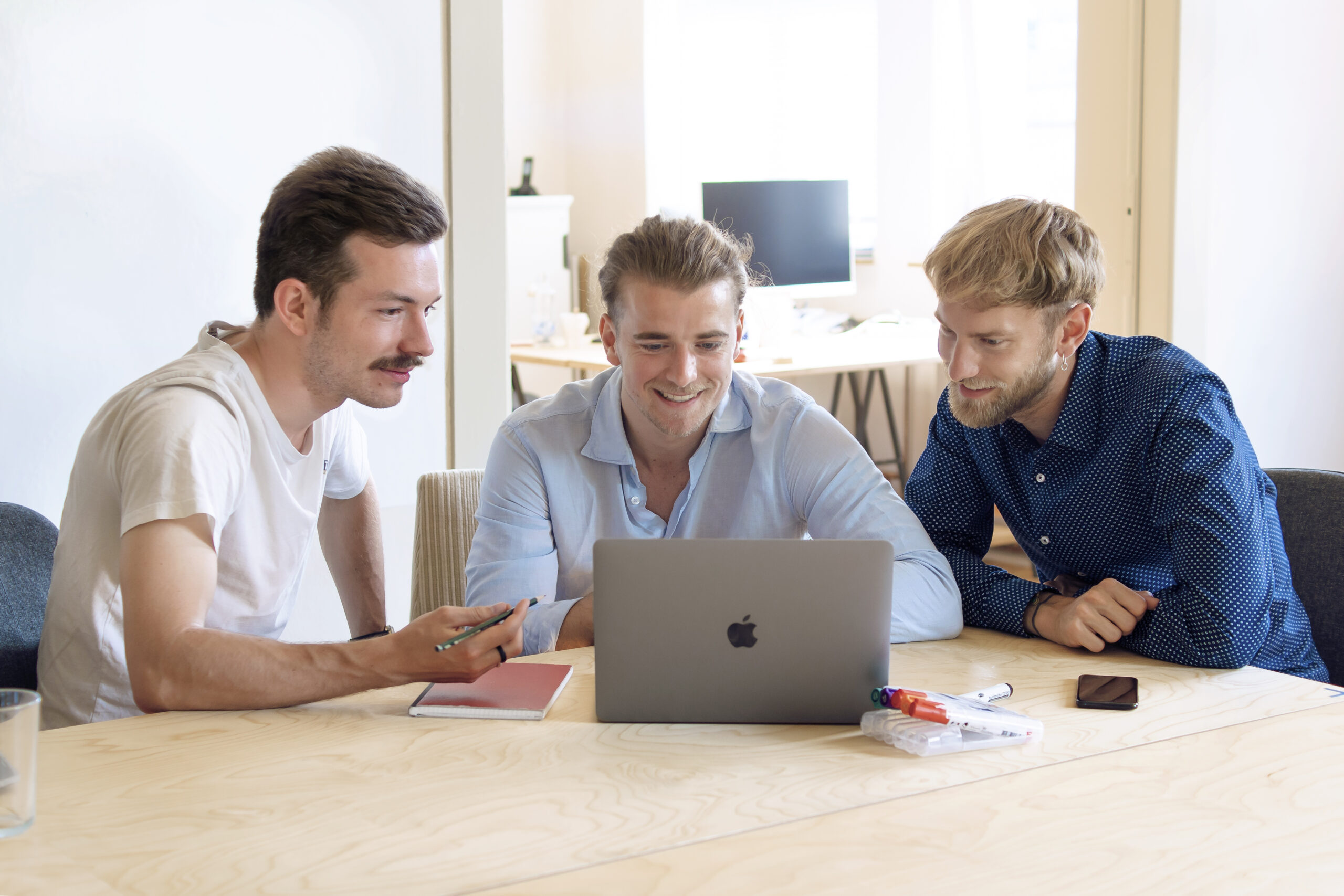 Drei Männer sitzen lächelnd um einen Laptop herum und diskutieren. Sie sitzen an einem Holztisch mit Notizbüchern, Stiften und einem Smartphone in der Nähe. Es scheint, als wären sie in einem hellen Büroraum der Hochschule Coburg.