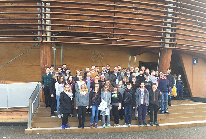 Eine Gruppe leger gekleideter Menschen posiert für ein Foto vor dem geschwungenen Holzeingang der Hochschule Coburg, zu dem eine Treppe hinaufführt. Sie stehen auf einer Betonfläche und halten den gemeinsamen Moment fest.