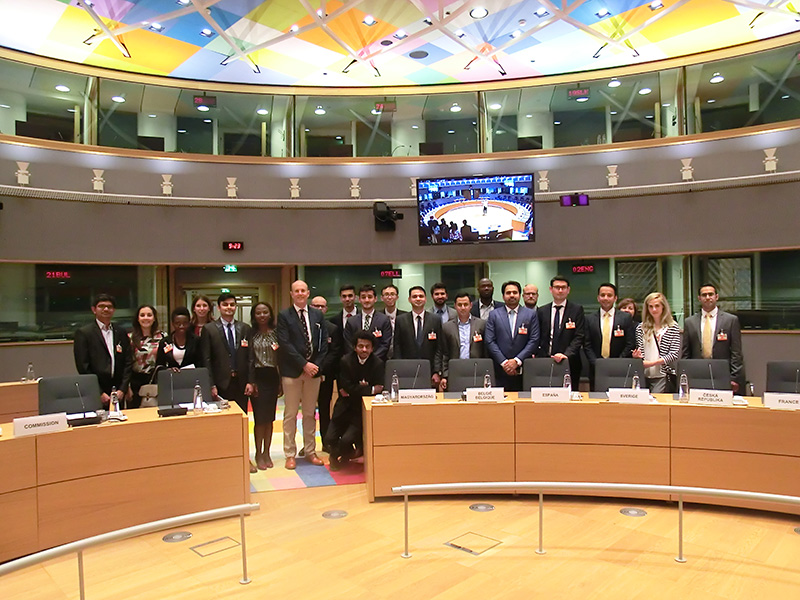 Eine Gruppe von Menschen in formeller Kleidung versammelt sich in einem Konferenzraum mit geschwungenen Schreibtischen und Namensschildern für Länder. Die bunte Decke spiegelt die dynamische Atmosphäre wider, während auf einem großen Bildschirm ein Meeting übertragen wird. Einige Teilnehmer, darunter Vertreter der Hochschule Coburg, tragen Namensschilder.