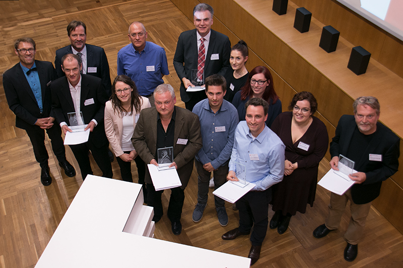 Eine Gruppe von Menschen steht auf einem Holzboden in der Hochschule Coburg und hält stolz Zertifikate und Auszeichnungen in den Händen. Sie blicken in die Kamera, manche lächeln. Im Raum stehen eine große Leinwand und ein Podium, was auf eine formelle Veranstaltung oder Zeremonie schließen lässt.