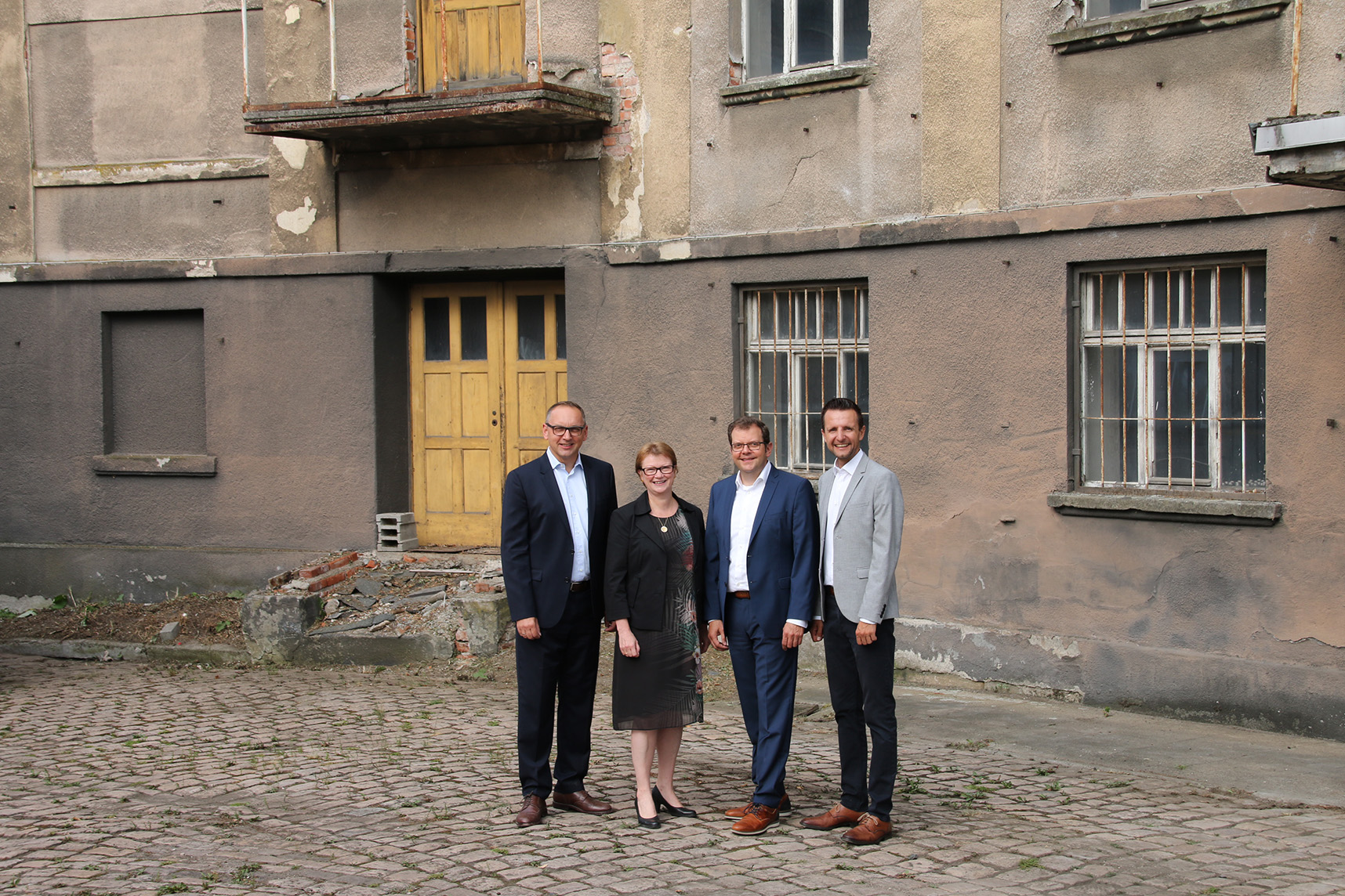 Vier Personen stehen vor einem alten, abgenutzten Gebäude mit einer gelben Tür, die an die Architektur der Hochschule Coburg erinnert. Sie sind in Geschäftskleidung gekleidet und stehen auf einem Kopfsteinpflaster. Die abblätternde Farbe und die vergitterten Fenster lassen darauf schließen, dass das Gebäude verlassen oder in schlechtem Zustand ist.