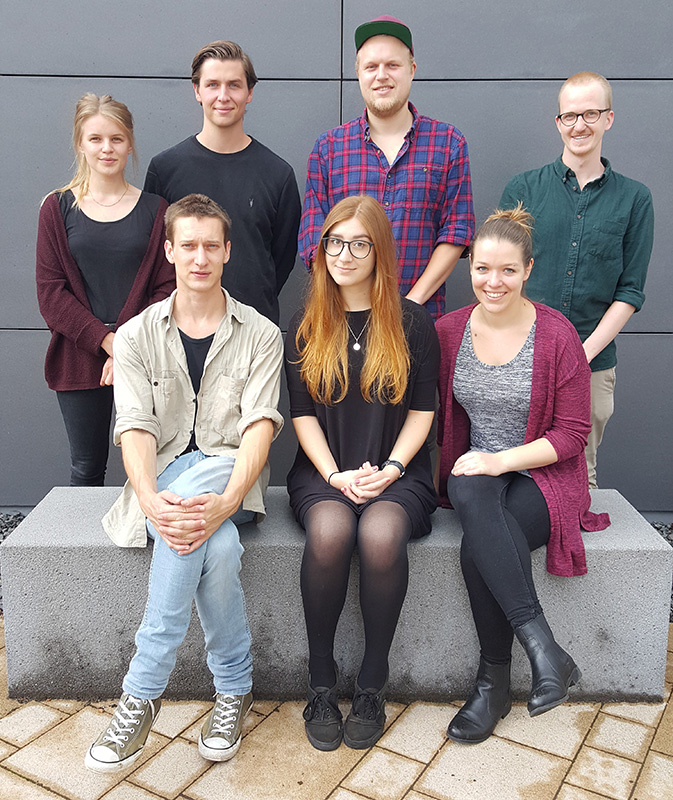 Eine Gruppe von sieben Personen, leger gekleidet, lächelnd und posierend vor einer grauen Wand der Hochschule Coburg. Sie sitzen in zwei Reihen auf einer Steinbank und schaffen so eine lebendige und doch entspannte Atmosphäre.