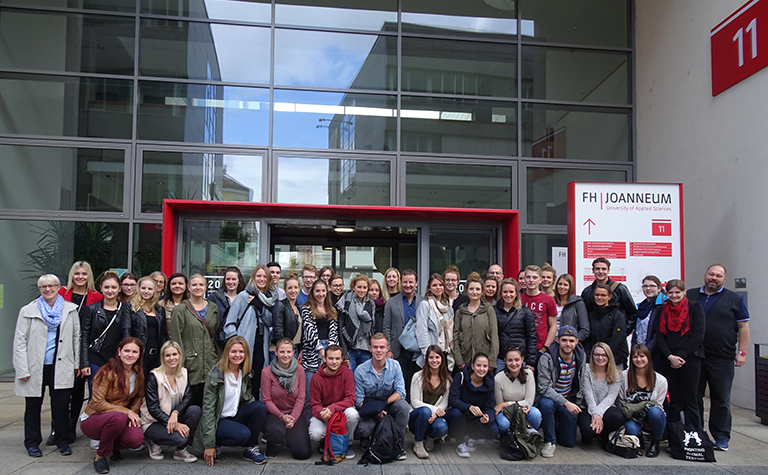 Eine Gruppe von Menschen posiert für ein Foto vor dem modernen Eingang mit Glasfenstern. Auf dem Schild steht „FH Joanneum“ und erinnert an den lebendigen Campusgeist der Hochschule Coburg. Die Gruppe besteht aus einer Mischung von Männern und Frauen, leger gekleidet, und fängt einen Moment gemeinsamer akademischer Begeisterung ein.