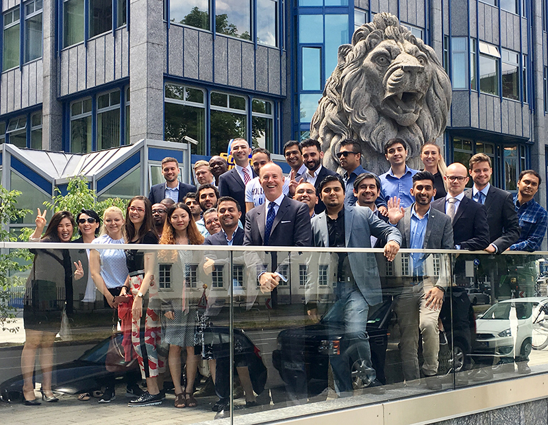 Eine bunt gemischte Gruppe von Menschen in legerer Business-Kleidung posiert gemeinsam vor einer großen Löwenstatue vor dem modernen Gebäude der Hochschule Coburg. Sie lächeln und gestikulieren fröhlich in Richtung Kamera.