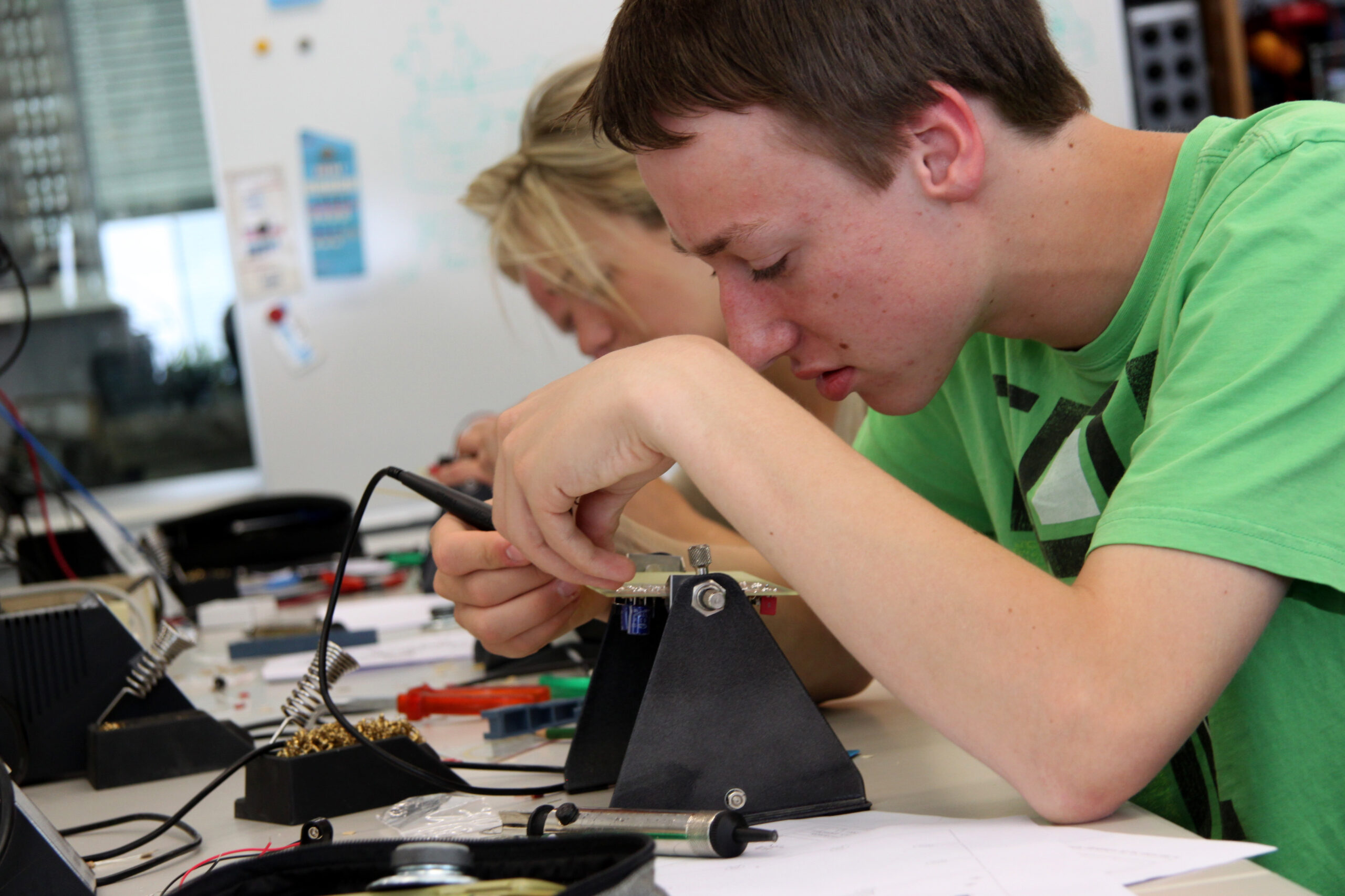 Ein junger Mann im grünen Hemd lötet konzentriert an seinem Schreibtisch ein kleines elektronisches Bauteil. Überall liegen Werkzeuge und Kabel herum, was an die innovative Atmosphäre der Hochschule Coburg erinnert. Im Hintergrund widmet sich eine Frau mit Präzision und Hingabe einer ähnlichen Aufgabe.