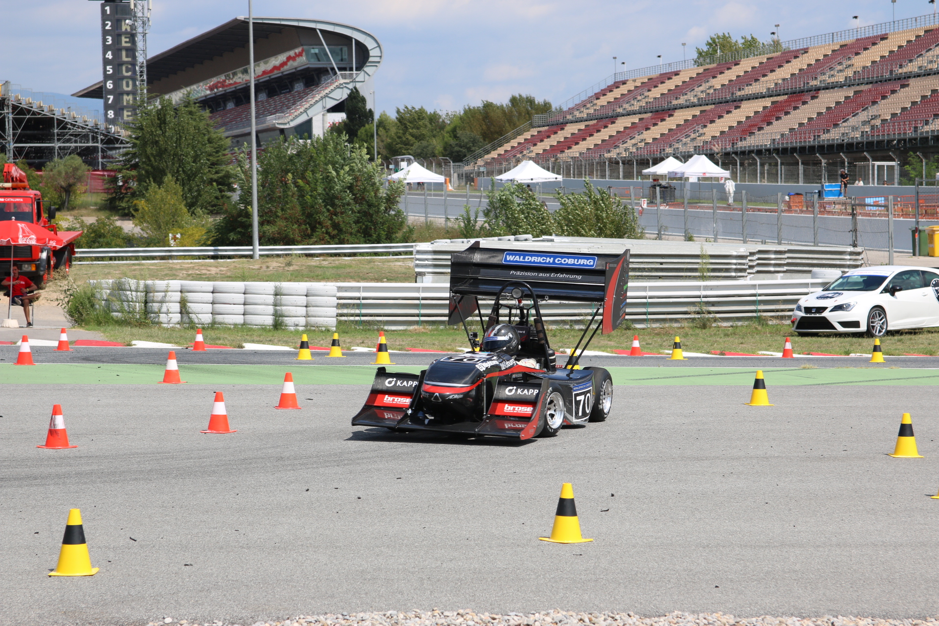 Ein schwarzer Rennwagen mit der Nummer 70 manövriert gekonnt um orangefarbene Pylonen bei einer Motorsportveranstaltung der Hochschule Coburg. In der Nähe parkt ein weißer Wagen, und unter dem teilweise bewölkten Himmel sind die Tribünen zu sehen.