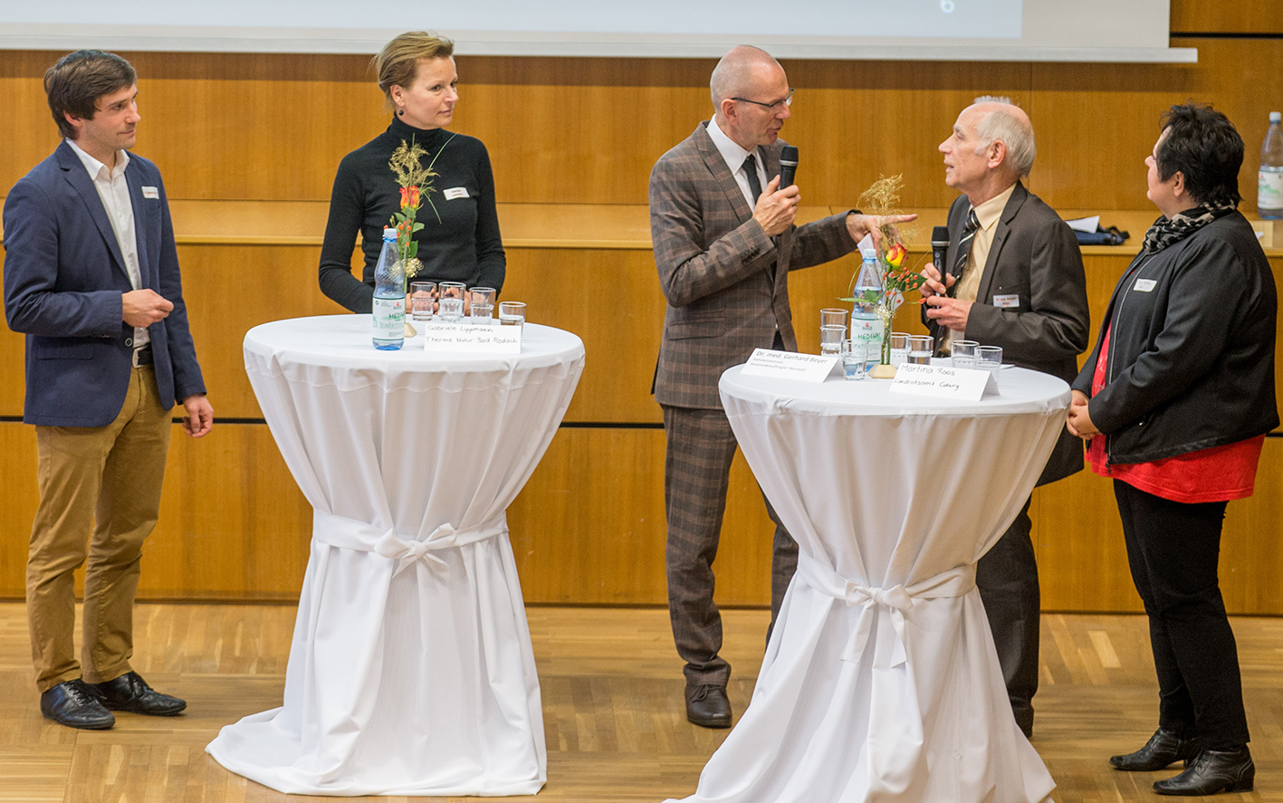 Fünf Personen stehen um zwei Stehtische in einem Konferenzraum der Hochschule Coburg. Sie scheinen in eine Diskussion vertieft zu sein. Eine Person hält ein Mikrofon. Jeder Tisch ist mit einem weißen Tuch bedeckt und steht mit Wasserflaschen und Gläsern darauf.