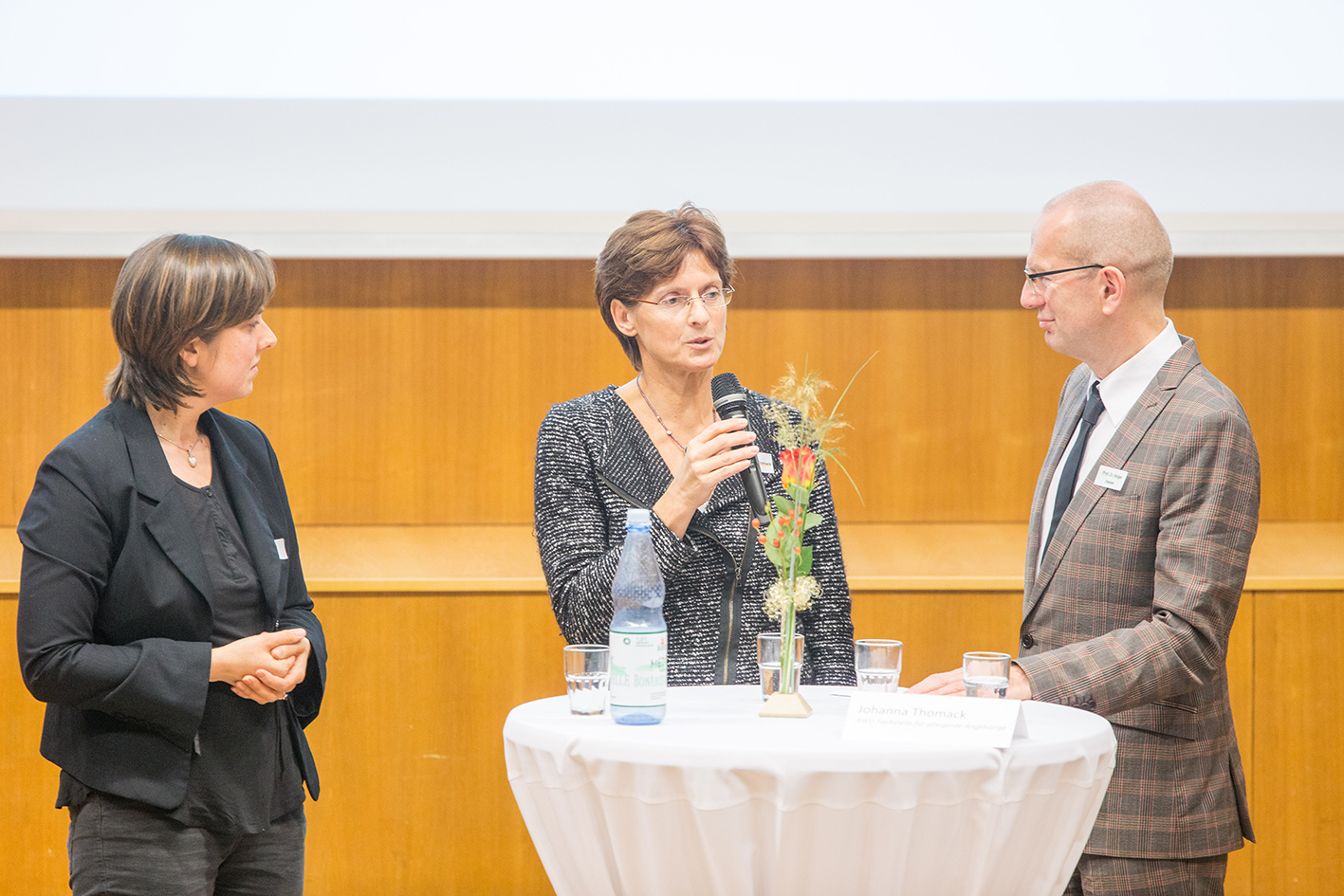 Drei Personen stehen bei der Veranstaltung der Hochschule Coburg um einen runden Tisch mit weißer Tischdecke. In der Mitte spricht eine Frau in ein Mikrofon, links flankiert von einer Frau und rechts von einem Mann. Auf dem Tisch stehen Wasserflaschen und ein kleines Blumenarrangement.