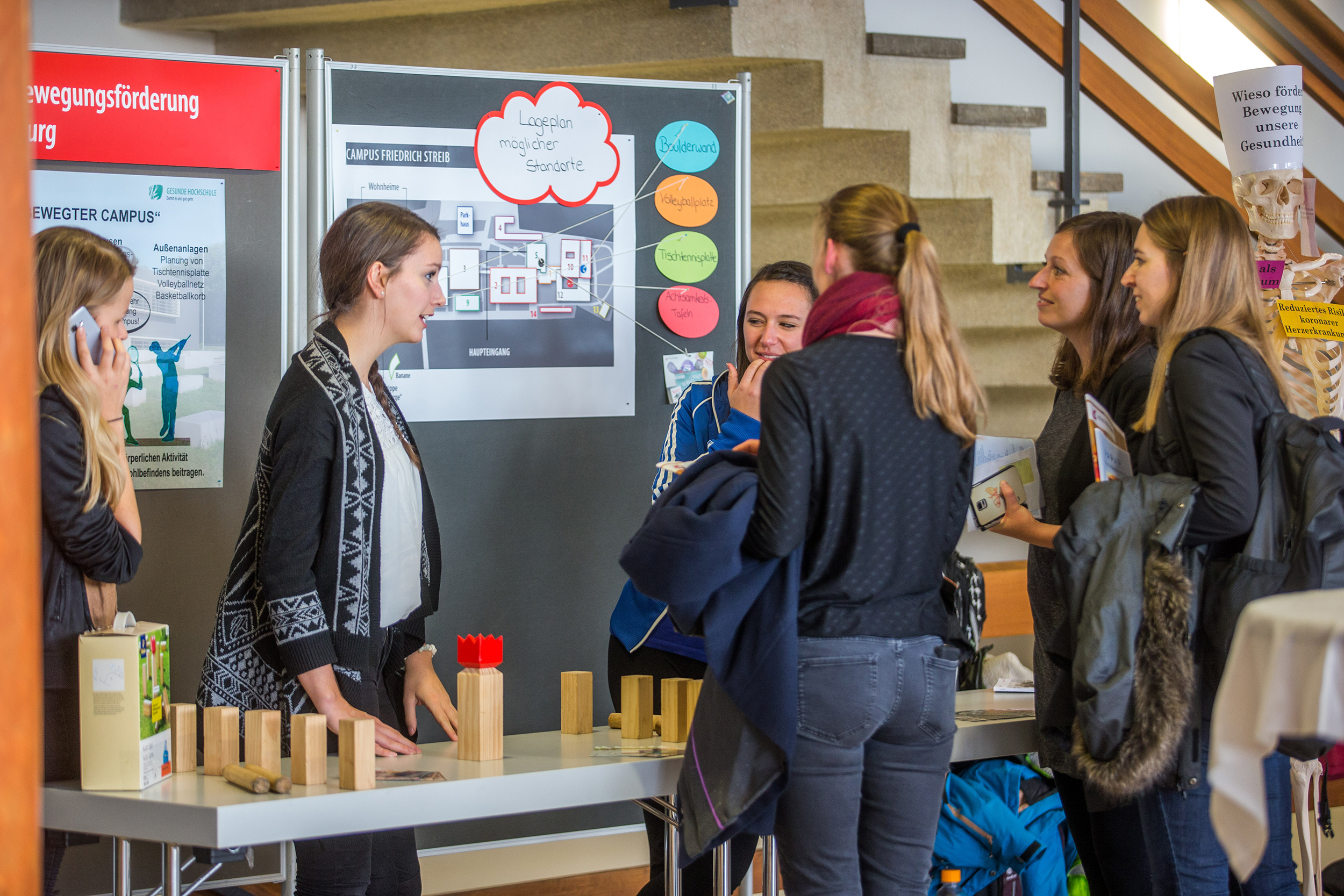 Eine Gruppe von Frauen versammelte sich an einem Tischausstellungsstand der Hochschule Coburg und unterhielt sich zwischen Informationstafeln. Holzklötze und Materialien, die über den Tisch verteilt sind, wecken das Interesse, während nahe gelegene Treppen und teilweise sichtbare Poster auf eine lebendige akademische Umgebung hinweisen.