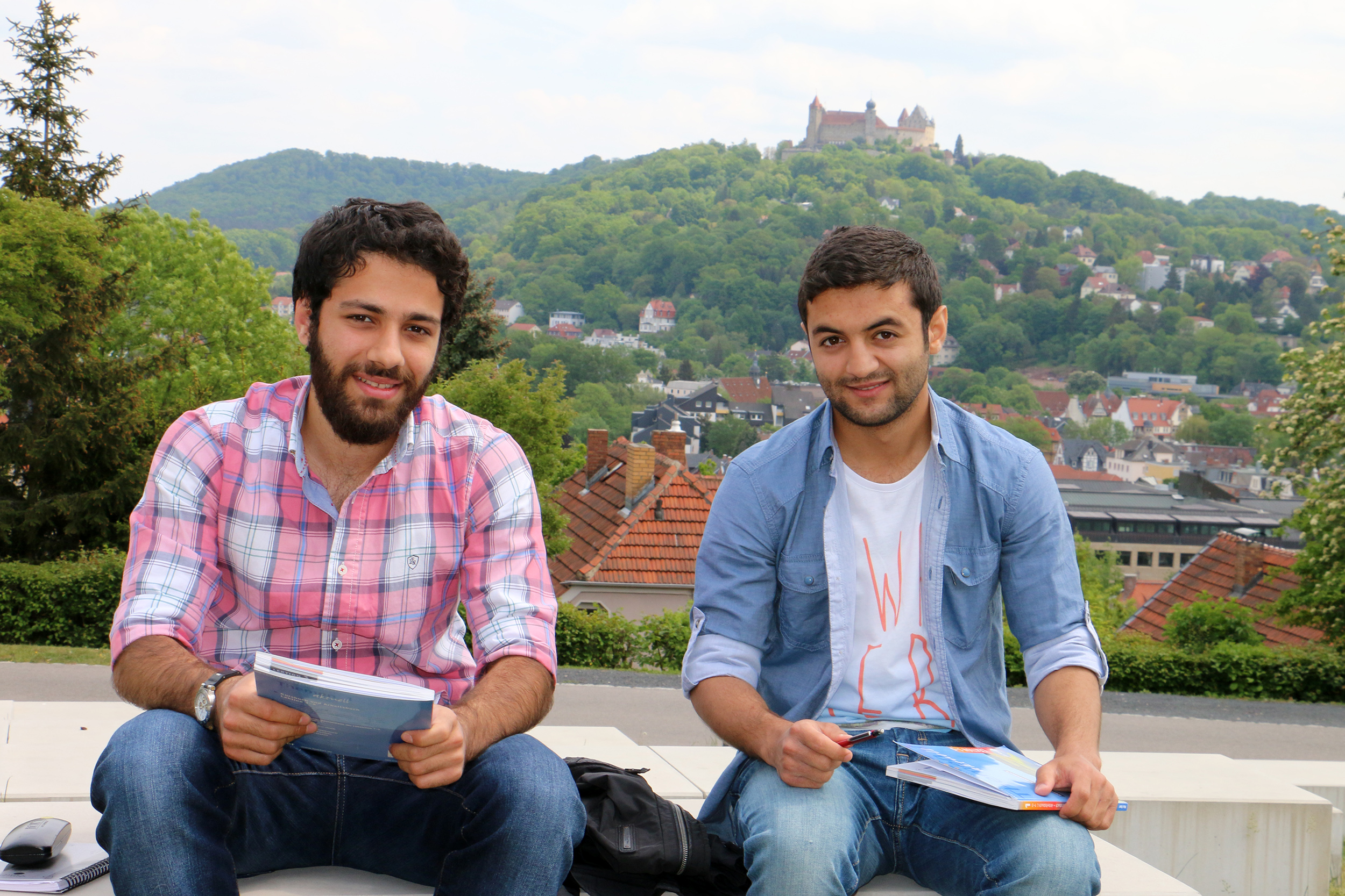 Zwei junge Männer sitzen mit Notizbüchern und Stiften in den Händen auf einer Bank im Freien an der Hochschule Coburg. Zu sehen ist eine malerische Landschaft mit Hügeln, Bäumen und einer Burg im Hintergrund.