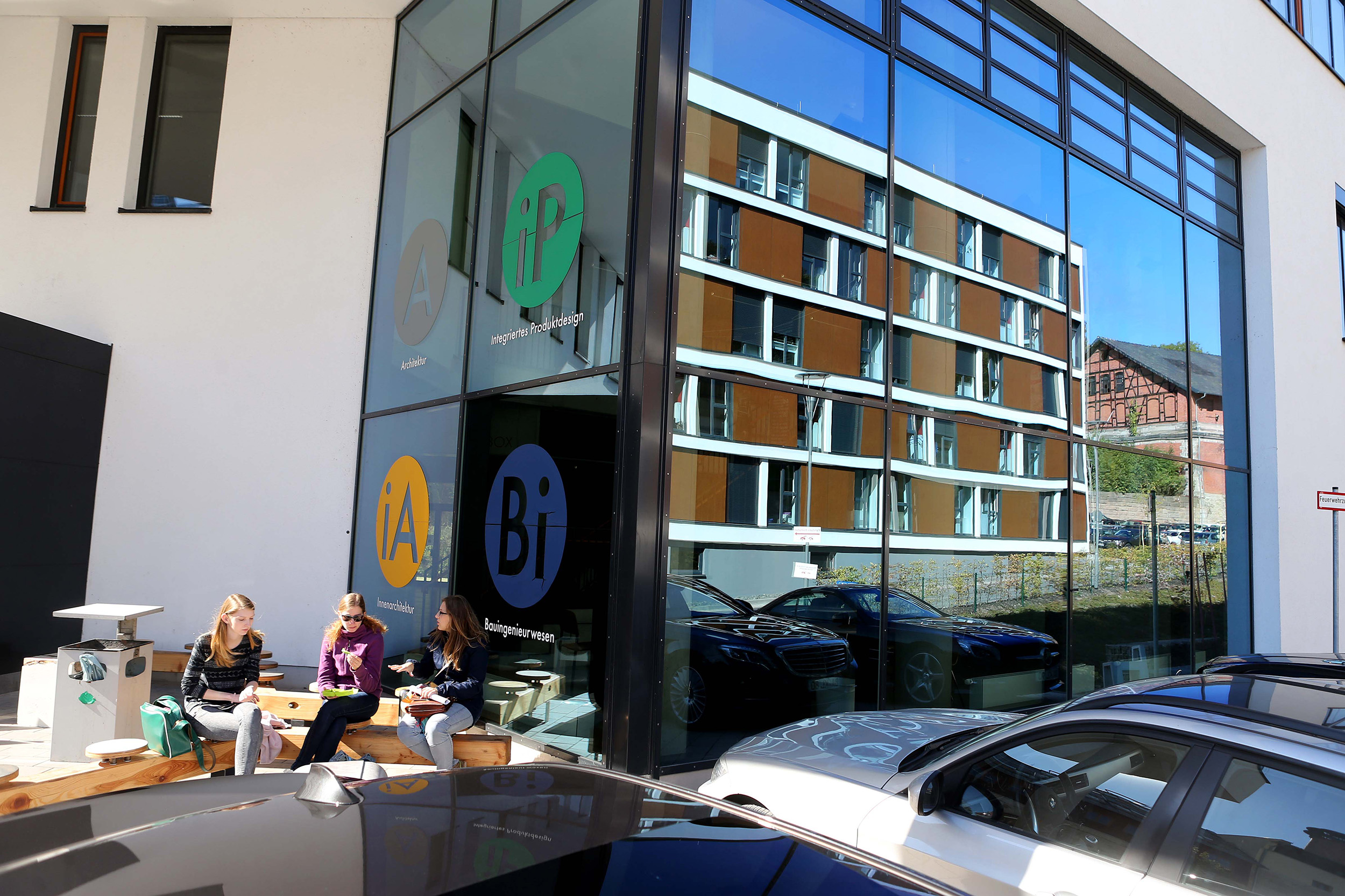 Drei Menschen sitzen auf Bänken vor einem modernen Gebäude mit großen Fenstern der Hochschule Coburg. Die Spiegelung zeigt ein weiteres Gebäude und eine hügelige Landschaft. Auf dem Glas sind runde Logos mit Buchstaben zu sehen. In der Nähe parken mehrere Autos, die zur lebendigen Atmosphäre des Campus beitragen.