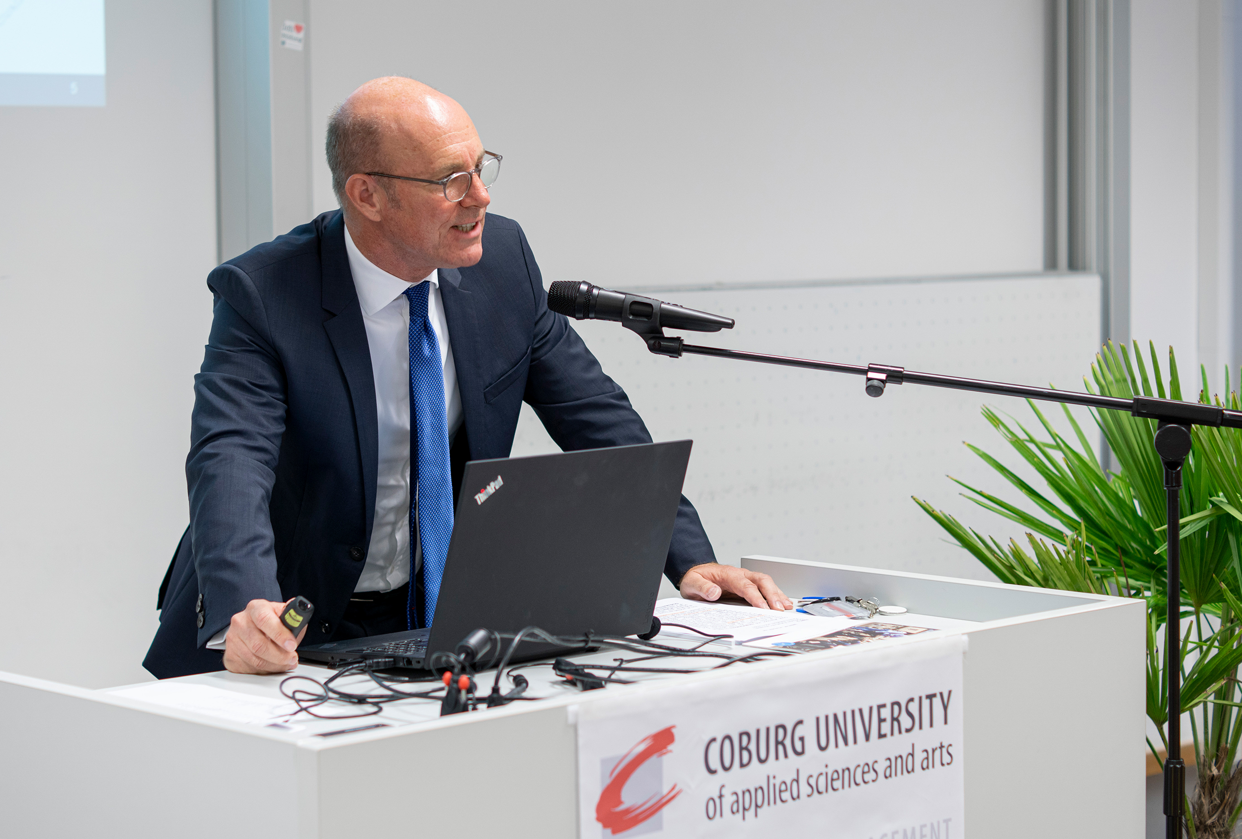 Ein Mann im Anzug hält an einem Podium mit Laptop und Mikrofon eine Präsentation in einem Raum. Über dem Pult steht auf einem Banner „Hochschule Coburg University of Applied Sciences and Arts“.