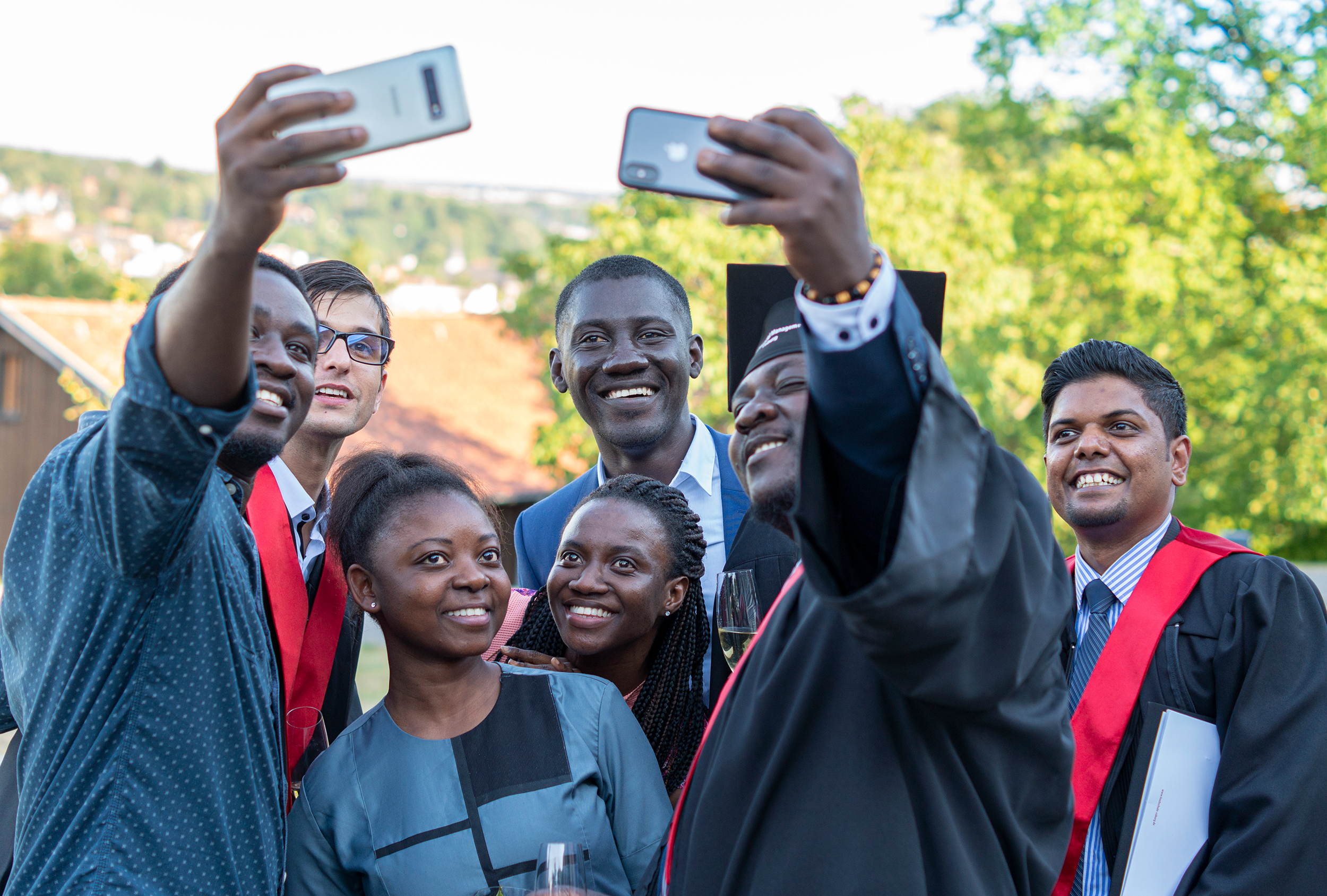 Eine Gruppe lächelnder Absolventen der Hochschule Coburg in Talaren und Kappen macht Selfies im Freien. Sie stehen dicht beieinander und halten den freudigen Moment fest. Im Hintergrund sind Bäume und ein Gebäude zu sehen.