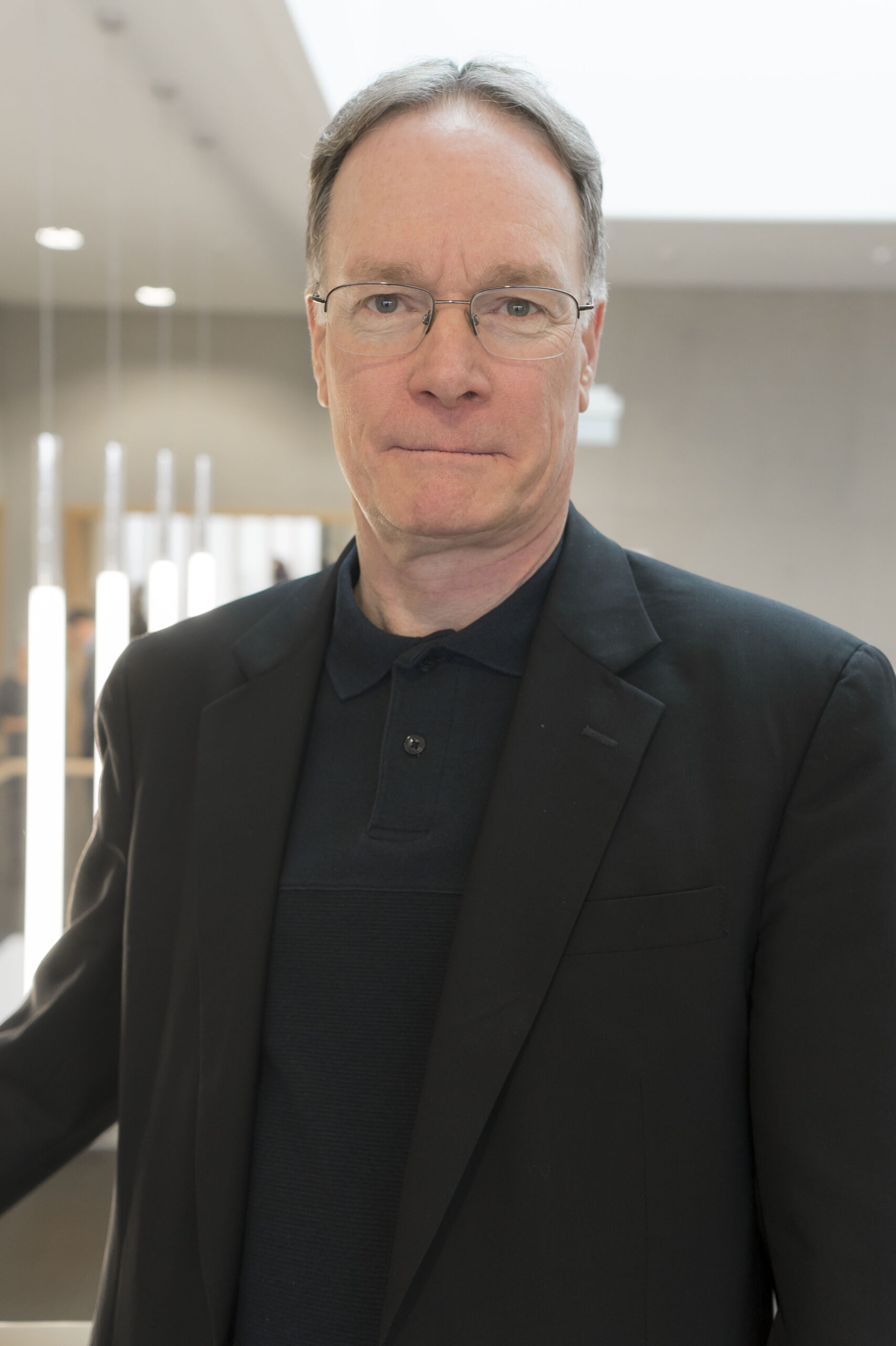 Ein Mann mit Brille, einer dunklen Jacke und einem Hemd mit Kragen steht im Innenbereich der Hochschule Coburg vor einem unscharfen Hintergrund mit vertikalen Lichtern.