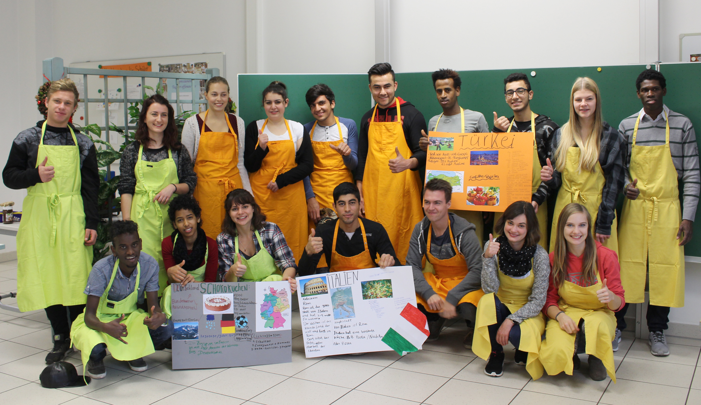 Eine Gruppe unterschiedlicher junger Leute in gelben Schürzen posiert in einem Klassenzimmer der Hochschule Coburg. Einige halten bunte Plakate mit den Themen Essen und Mahlzeiten, andere strecken den Daumen hoch. Im Hintergrund sind eine Tafel und farbenfrohe Klassenzimmerpflanzen zu sehen, die zur lebhaften Atmosphäre beitragen.