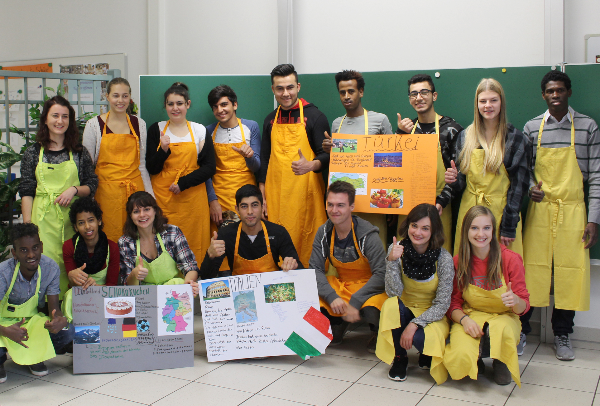 Eine bunt gemischte Gruppe von Studierenden in gelben Schürzen und Freizeitkleidung posiert in einem Hörsaal der Hochschule Coburg. Sie lächeln und strecken den Daumen hoch, während sie vor einer grünen Tafel bunte Plakate über verschiedene Länder, darunter die Türkei und Italien, in die Höhe halten.