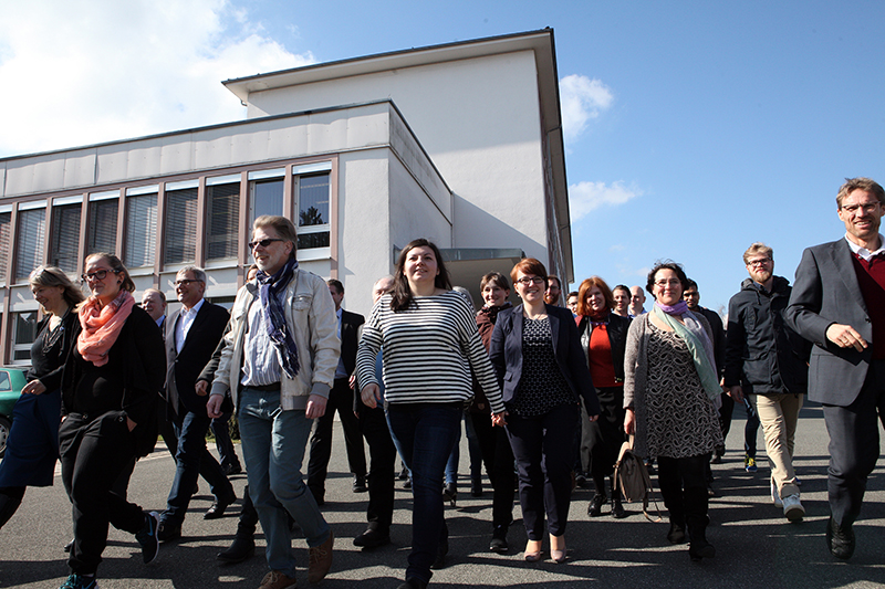 Eine Gruppe der Hochschule Coburg schlendert an einem sonnigen Tag gemeinsam vor einem modernen Gebäude umher. Sie wirken lächelnd und vertieft, im Hintergrund ein klarer blauer Himmel.