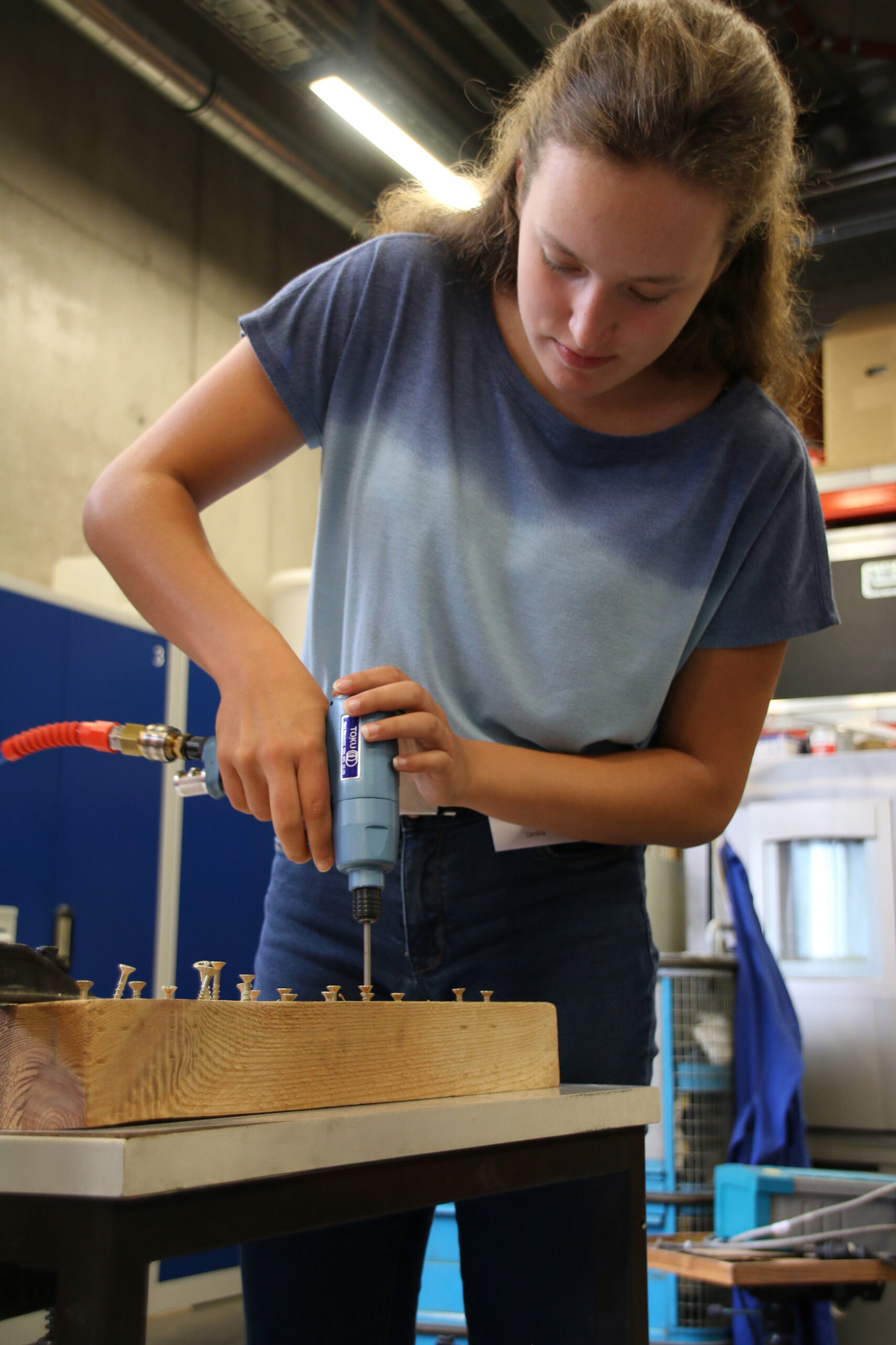 In einer geschäftigen Werkstatt bohrt eine Person in blauem Hemd und Jeans geschickt mit einem Elektrowerkzeug Schrauben in ein Holzbrett. Die Kulisse der Hochschule Coburg wird durch Industrieanlagen und Regale im Hintergrund akzentuiert.