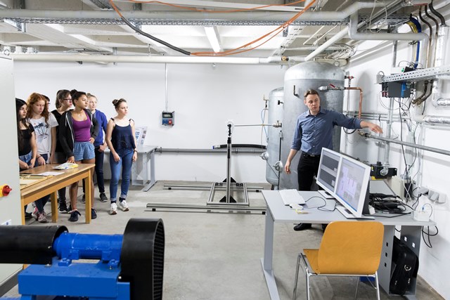 Ein Mann im blauen Hemd erklärt einer Gruppe von Studenten die Ausstattung eines Labors der Hochschule Coburg. Sie stehen neben einem Tisch mit Papieren, einem großen Monitor und Industriemaschinen in einem Raum mit freiliegenden Rohren und Kabeln.