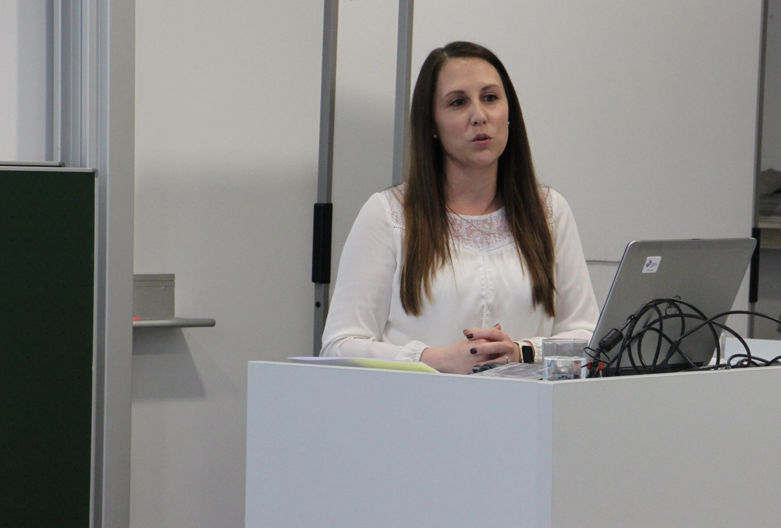 Eine Frau mit langen braunen Haaren spricht an einem Podium in der Hochschule Coburg. Sie trägt eine weiße Bluse und steht vor einem Laptop und einigen Kabeln. Hinter ihr sind ein Whiteboard und eine geschlossene Tür.