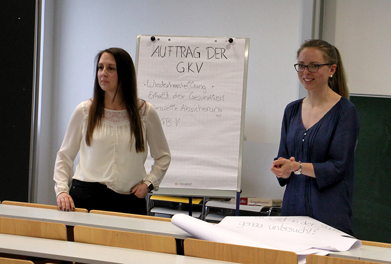 Zwei Frauen stehen vor einem Hörsaal der Hochschule Coburg. Eine schreibt mit Stichpunkten auf Deutsch „AUFTRAG DER GKV“ auf ein Flipchart, die andere lächelt neben ihr. Im Vordergrund stehen leere Tische, die Bühne für eine spannende Vorlesung.