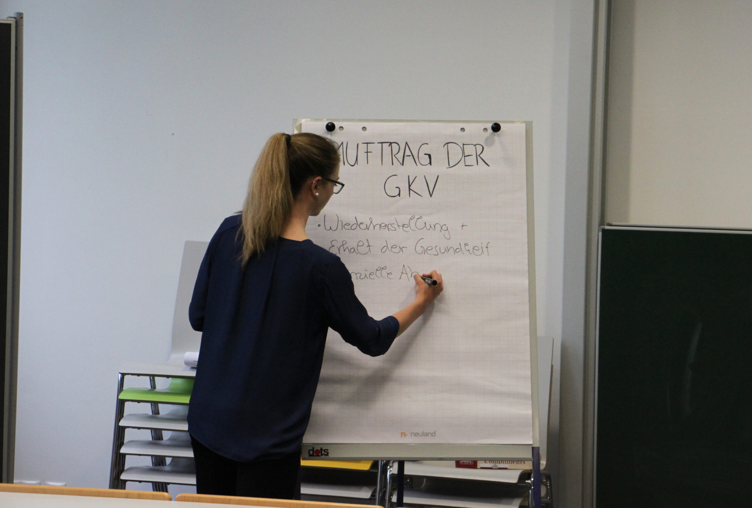 Eine Person steht vor einem Flipchart und schreibt auf Deutsch an der Hochschule Coburg. Der Text auf dem Flipchart enthält „AUFTRAG DER GKV“. Die Szene scheint ein Klassenzimmer oder ein Konferenzraum zu sein, im Hintergrund sind Regale zu sehen.