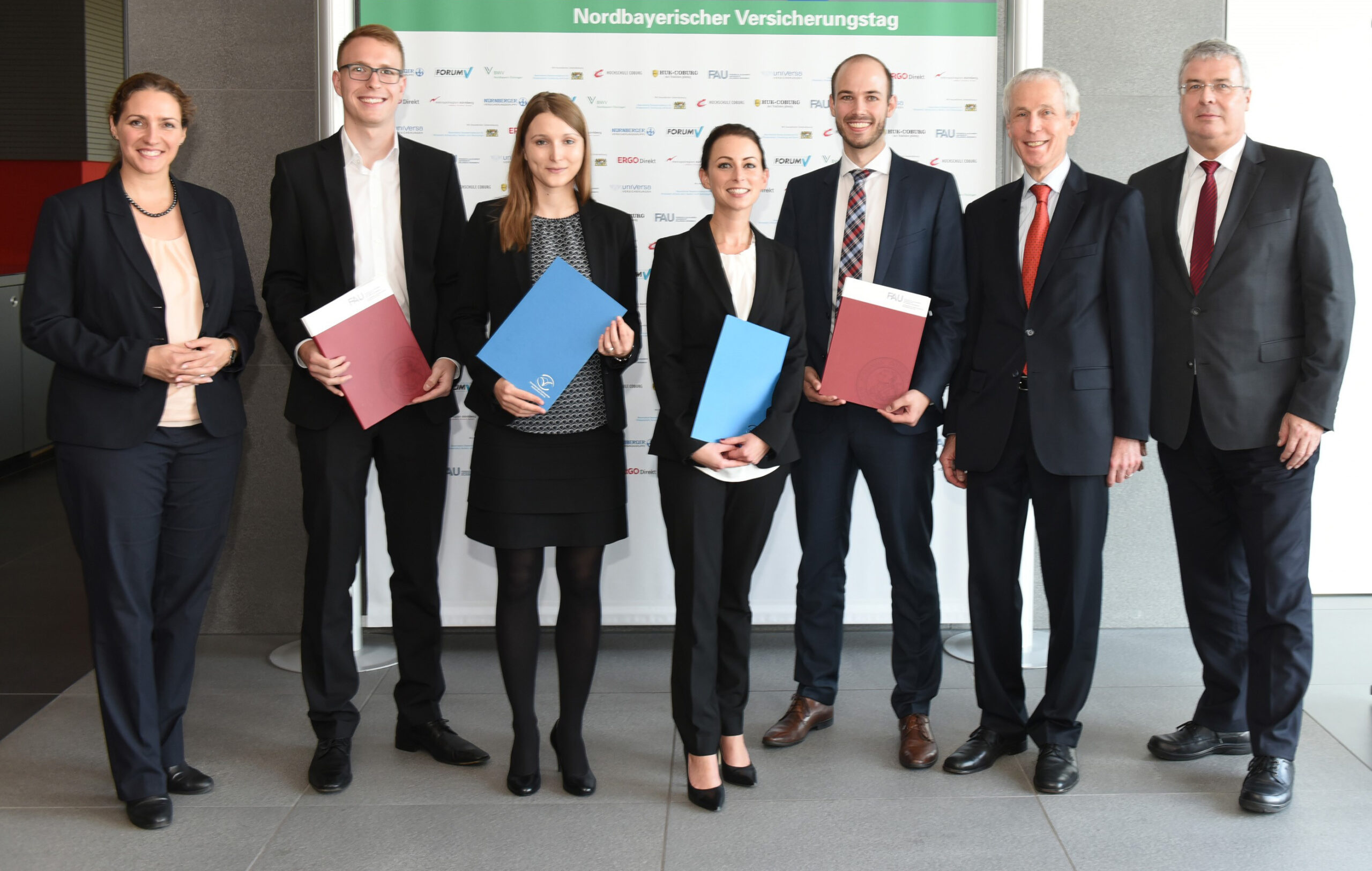 Eine Gruppe von sieben Personen der Hochschule Coburg, alle in formeller Kleidung, steht drinnen und lächelt für ein Foto. Drei Personen halten stolz Ordner in den Händen. Ein Hintergrund mit Logos und dem Text „Nordbayerischer Versicherungstag“ verleiht dem Moment eine offizielle Note.