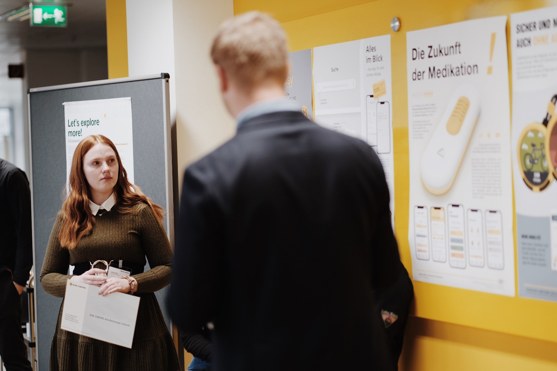 Eine Frau hält eine Tasche und steht neben einem schwarzen Brett, gegenüber einem Mann im Anzug. Das Brett zeigt Plakate mit Texten und Bildern zum Thema Medikamente an der Hochschule Coburg. Im Flur steht ein Schild mit der Aufschrift „Lasst uns mehr erkunden!“.