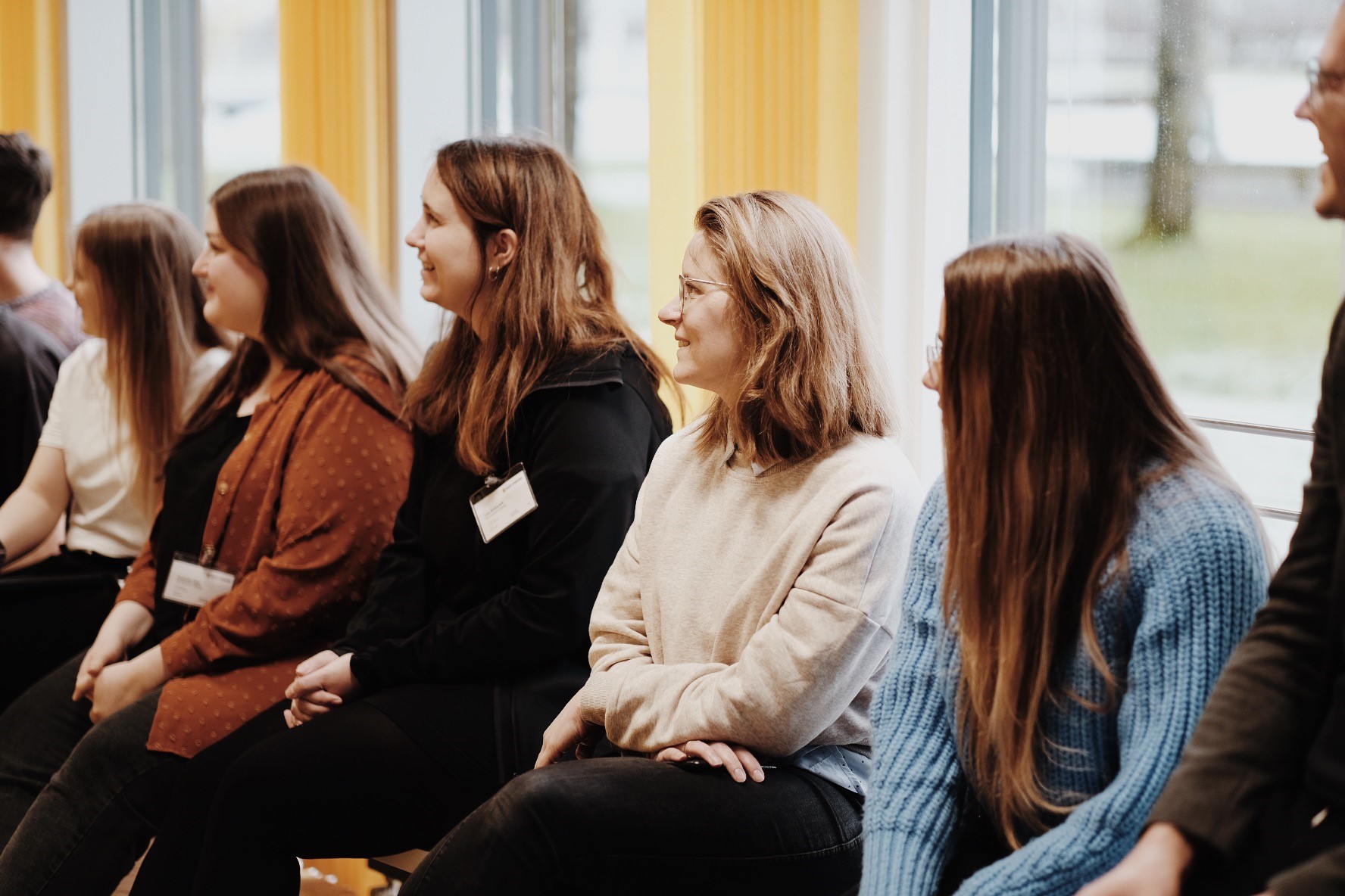 Eine Gruppe von Menschen sitzt auf Stühlen, wirkt engagiert und lächelnd während eines lockeren Treffens in Innenräumen an der Hochschule Coburg. Sie befinden sich in einem hell erleuchteten Raum mit großen Fenstern im Hintergrund.