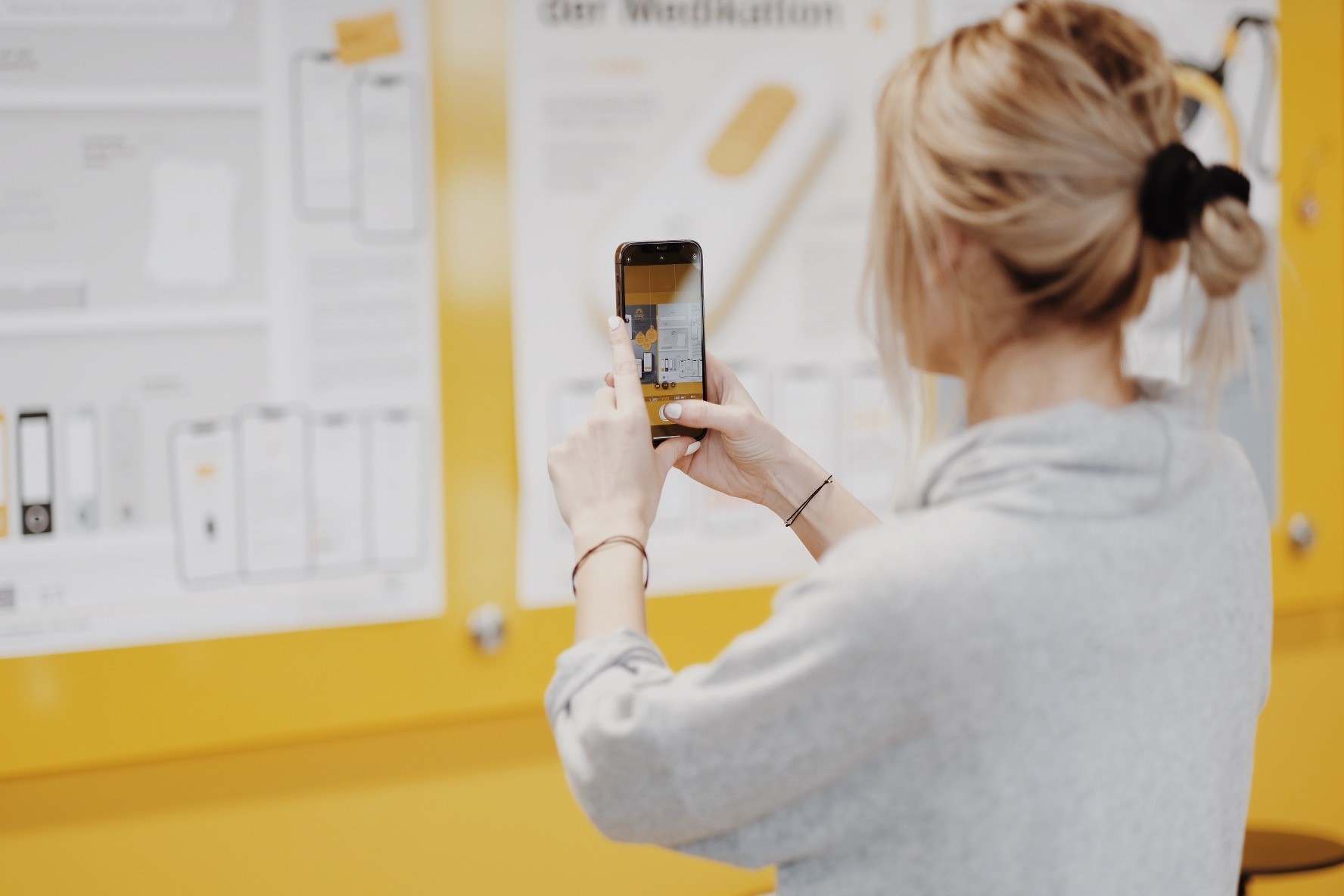 Eine Person mit blondem Haarknoten fotografiert mit einem Smartphone ein mit Diagrammen und Texten verziertes Wanddisplay der Hochschule Coburg. Der gelbe Hintergrund harmoniert mit dem hellen Langarmshirt der Person.