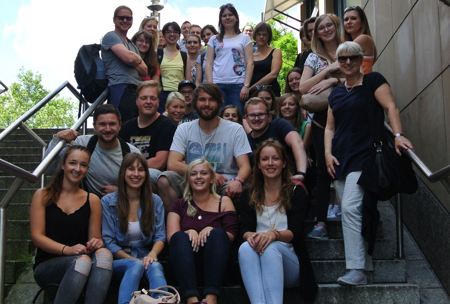 Eine Gruppe von Menschen sitzt und steht auf Außentreppen und posiert für ein Foto an der Hochschule Coburg. Sie sind leger gekleidet und scheinen Spaß zu haben, mit Bäumen und einem Gebäude im Hintergrund.