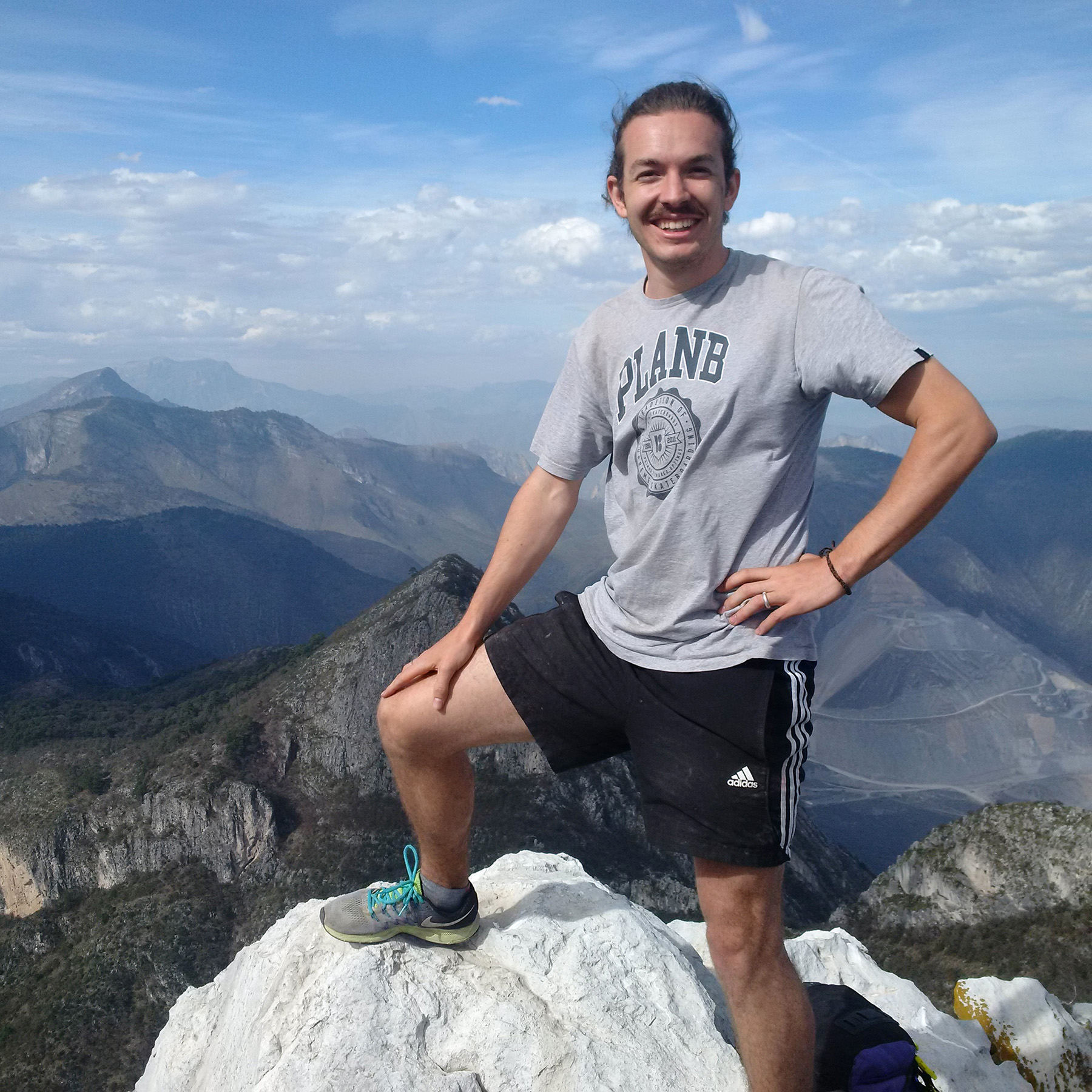 Eine Person steht auf einem Felsen auf einem Berggipfel und trägt stolz ein T-Shirt der Hochschule Coburg und schwarze Shorts. Der Himmel ist klar mit einigen Wolken und gibt den Blick auf die weiten Berge im Hintergrund frei – eine perfekte Mischung aus Natur und Wissenschaft.