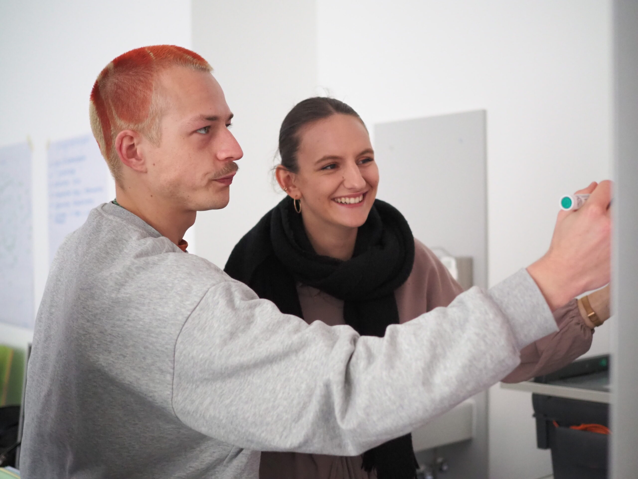 Zwei Personen arbeiten gemeinsam an einem Whiteboard der Hochschule Coburg. Die Person links mit den kurzen roten Haaren schreibt mit einem Marker, während die Person rechts lächelnd die Aktivität zu besprechen oder anzuleiten scheint.