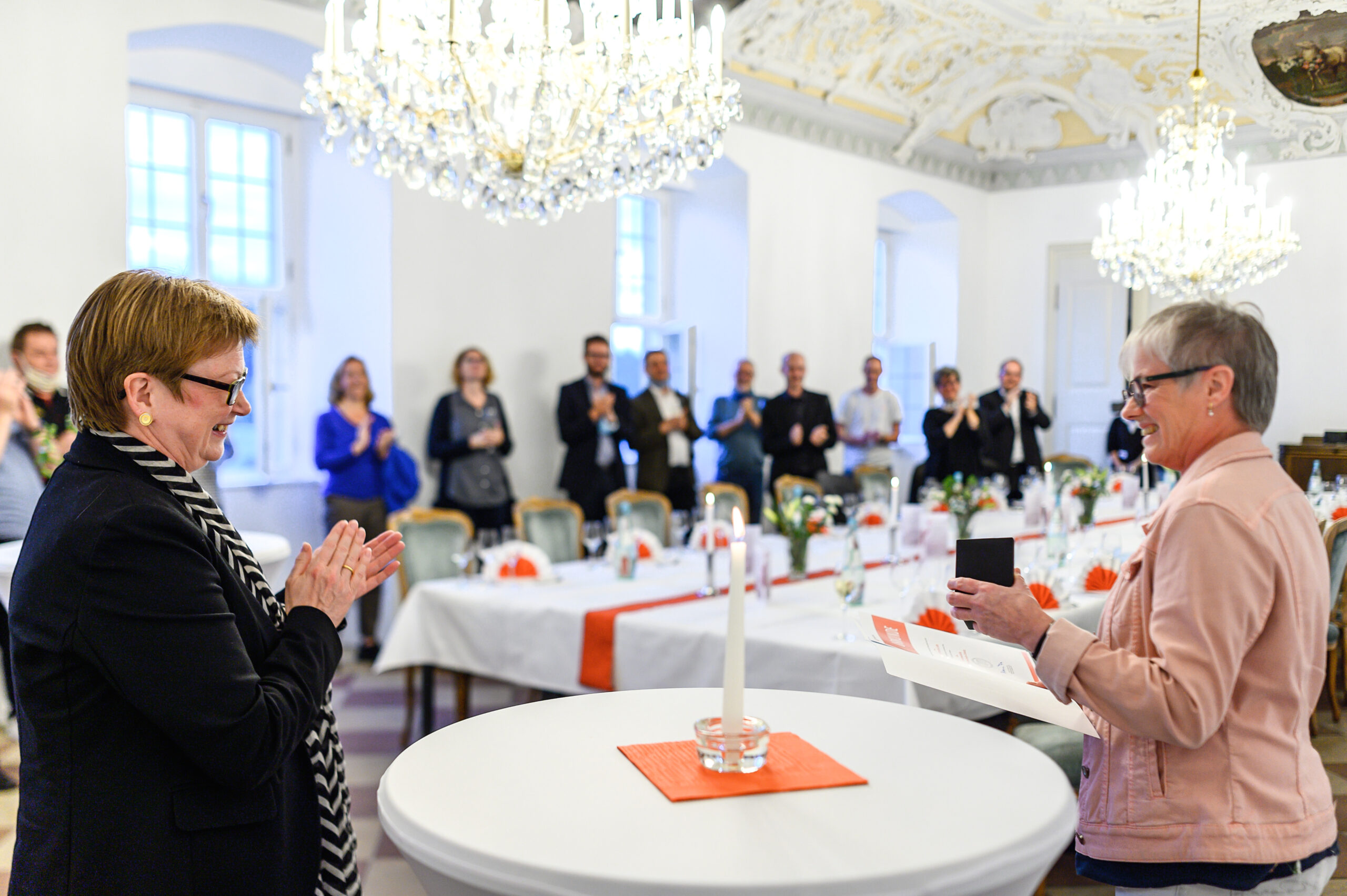 Zwei Frauen stehen an einem Banketttisch in einem eleganten, mit Kronleuchtern geschmückten Raum und feiern ein Ereignis. Eine Frau von der Hochschule Coburg applaudiert, während die andere eine Medaille in den Händen hält. Der mit Blumen und einer Kerze geschmückte Tisch bildet das Zentrum, während die Menschen im Hintergrund jubeln und klatschen.