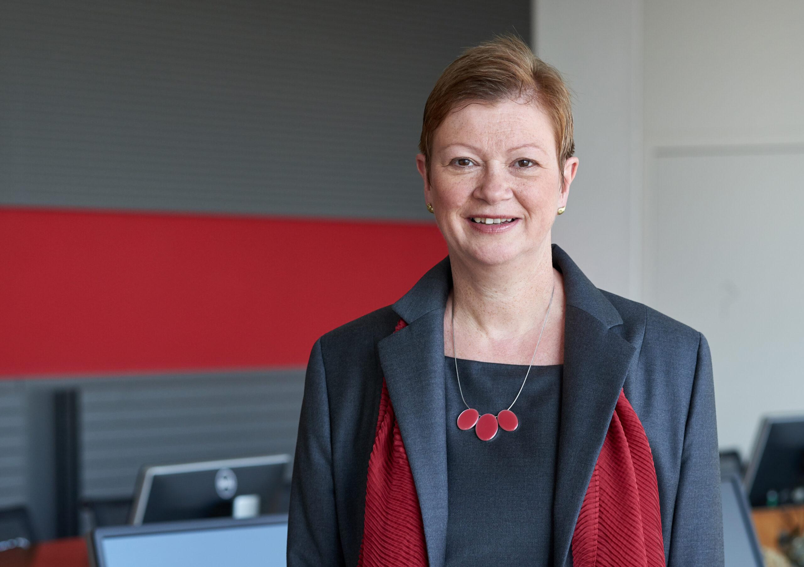 Eine Person mit kurzen Haaren, einem grauen Blazer und einer roten Halskette steht lächelnd in einem Büro der Hochschule Coburg. Im Hintergrund sind rot-graue Wände und Computerstationen zu sehen, die den akademischen Aspekt mit modernem Design perfekt verbinden.