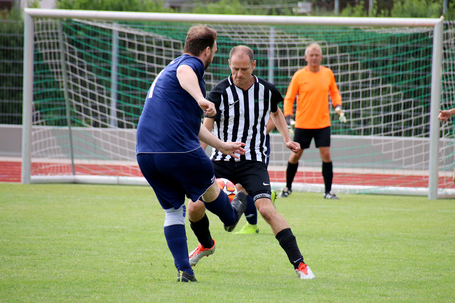 Auf einem Rasenplatz der Hochschule Coburg kämpfen zwei Fußballspieler um den Ball. Einer trägt ein blaues Trikot, der andere ein schwarz-weiß gestreiftes Trikot. Im Hintergrund sind ein Torwart in einem orangefarbenen Trikot und ein weiterer Spieler in der Nähe des Tores zu sehen.