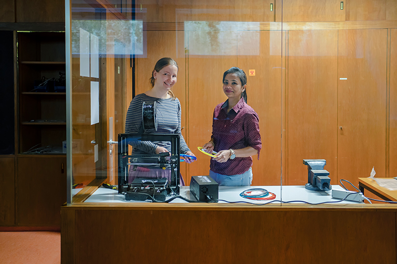 Zwei Frauen stehen lächelnd in einer Werkstatt der Hochschule Coburg hinter einer Glasscheibe. Eine hält ein Werkzeug in der Hand, während ein 3D-Drucker, ein Stapel Kabel und andere Geräte die hölzerne Werkbank vor ihnen überladen. Regale und Schränke säumen ordentlich die holzgetäfelten Wände.