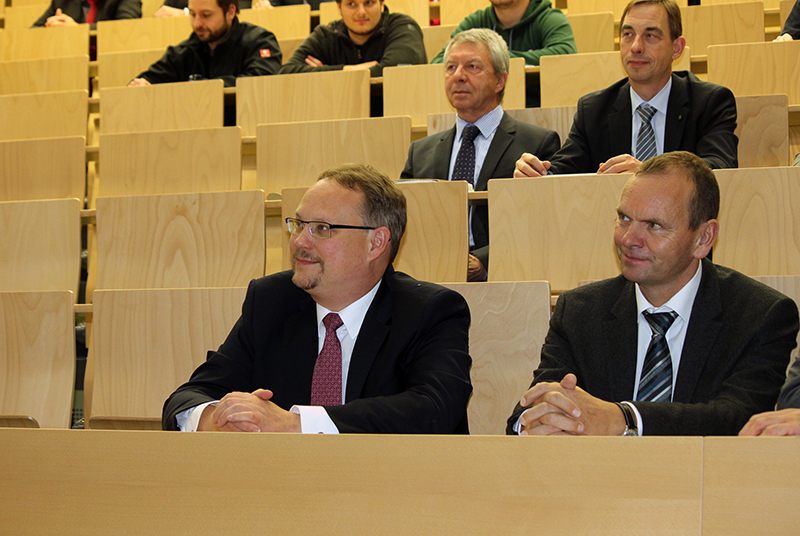 In einem Hörsaal der Hochschule Coburg mit Holzsitzen sitzt eine Gruppe von Männern in Anzügen aufmerksam. Der Fokus liegt auf zwei Männern in der ersten Reihe, die beide lächeln und engagiert sind. Im Hintergrund sind weitere Männer zu sehen, die der Diskussion aufmerksam zuhören.