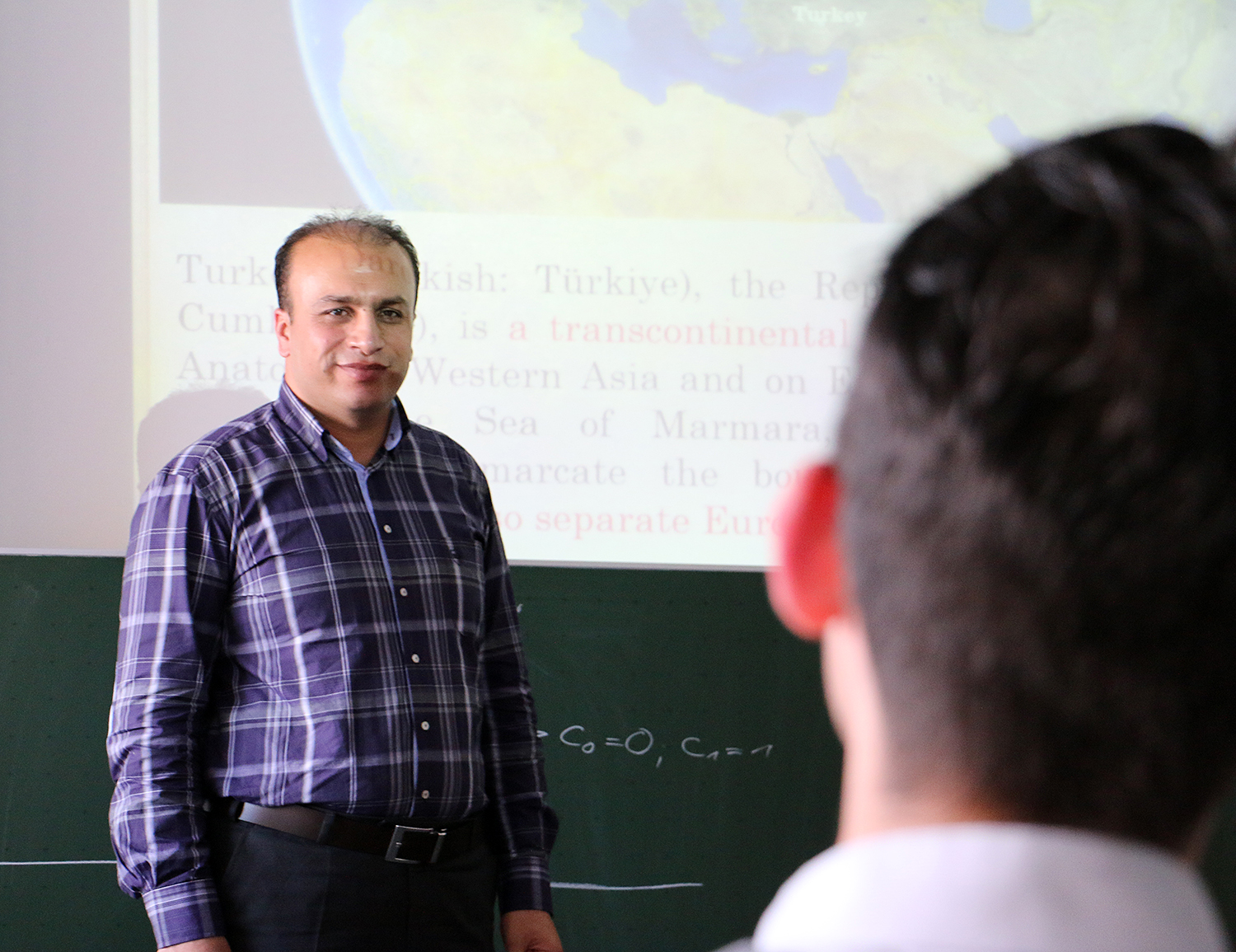 Ein Mann im karierten Hemd steht vor einer projizierten Präsentation an der Hochschule Coburg und spricht zu einer Gruppe von Studierenden. Auf der Leinwand werden eine Weltkarte und Informationen über die Türkei angezeigt. Im Vordergrund sitzt ein Student, unscharf, seine Aufmerksamkeit ist auf die Vorlesung gerichtet.