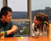 A man and a woman sit at a table by a window, engaged in conversation. On the table, marked with Hochschule Coburg's emblem, rests a bottle, a pad, and a yellow paper bag. Outside the window, a blurred hillside landscape is visible.