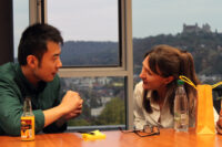 A man and a woman sit at a table by a window, engaged in conversation. On the table, marked with Hochschule Coburg's emblem, rests a bottle, a pad, and a yellow paper bag. Outside the window, a blurred hillside landscape is visible.