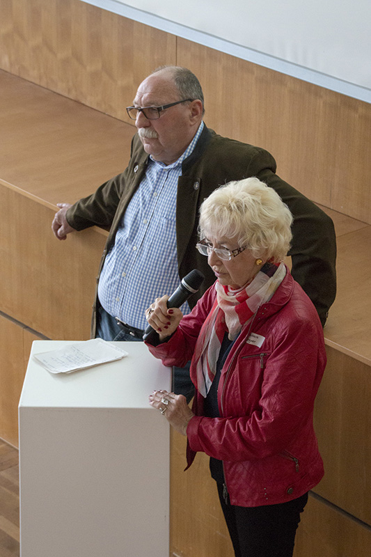 Eine ältere Frau in einer roten Jacke spricht in ein Mikrofon und wendet sich an ein Publikum an der Hochschule Coburg. Ein älterer Mann steht daneben und lehnt sich an ein Podium. Sie scheinen sich in einem hölzernen Hörsaal oder Konferenzraum zu befinden, was zur wissenschaftlichen Atmosphäre der Veranstaltung beiträgt.