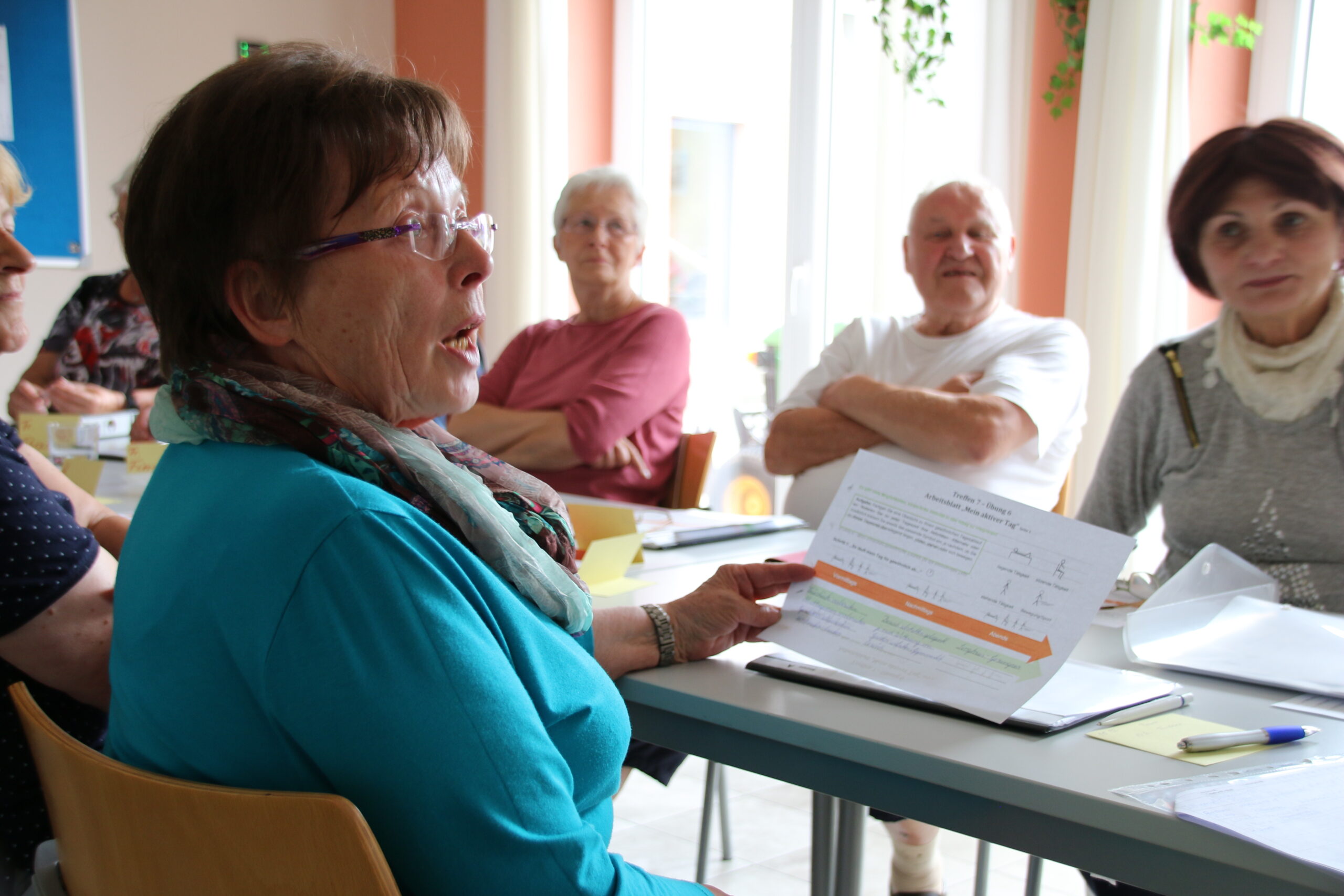 Eine Gruppe älterer Erwachsener sitzt in einem hellen Raum der Hochschule Coburg um einen Tisch. Eine Frau in blauem Hemd und Schal hält ein Dokument in der Hand und spricht zu der Gruppe. Die anderen hören aufmerksam zu. Auf dem Tisch liegen Papiere und Stifte.