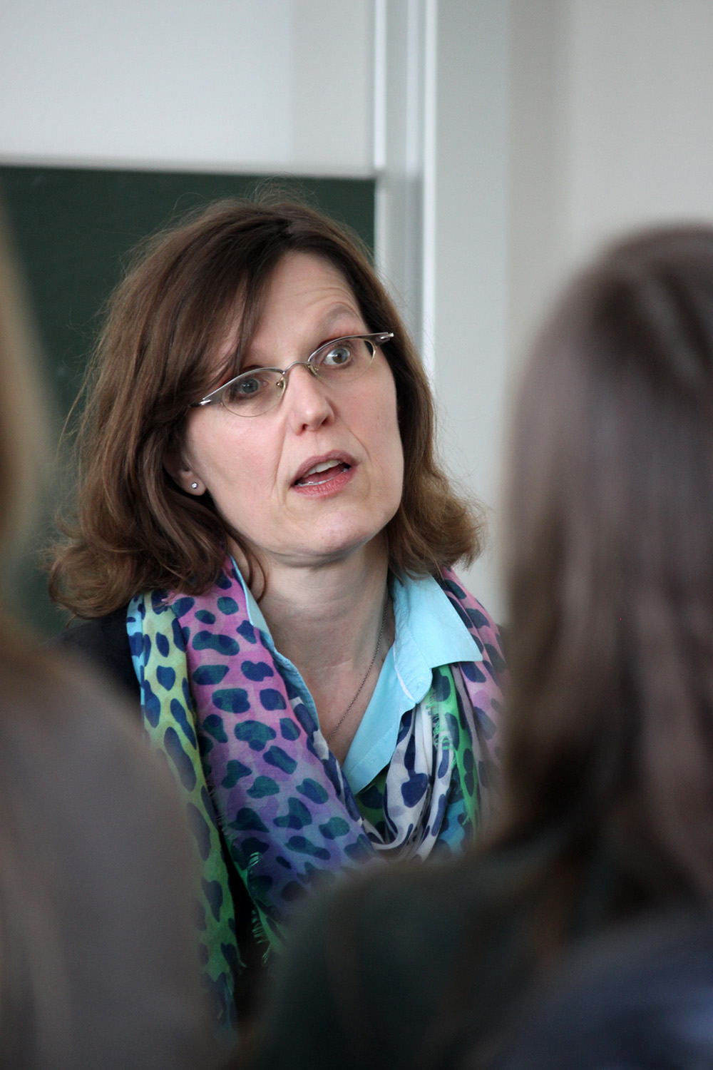 Eine Frau mit Brille und schulterlangem braunem Haar, einem bunten Schal und einem blauen Hemd bekleidet, steht vor einem Seminar der Hochschule Coburg. Sie scheint leidenschaftlich zu sprechen, während mehrere Teilnehmer im Vordergrund, leicht unscharf, gespannt ihren Ausführungen lauschen.