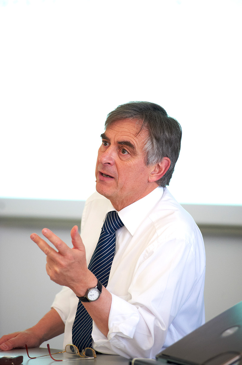 Ein Mann mit grauem Haar, weißem Hemd und blau gestreifter Krawatte sitzt an seinem Schreibtisch an der Hochschule Coburg und gestikuliert. Vor ihm steht ein Laptop, im Hintergrund ist ein Whiteboard zu sehen, das auf eine spannende akademische Diskussion hindeutet.