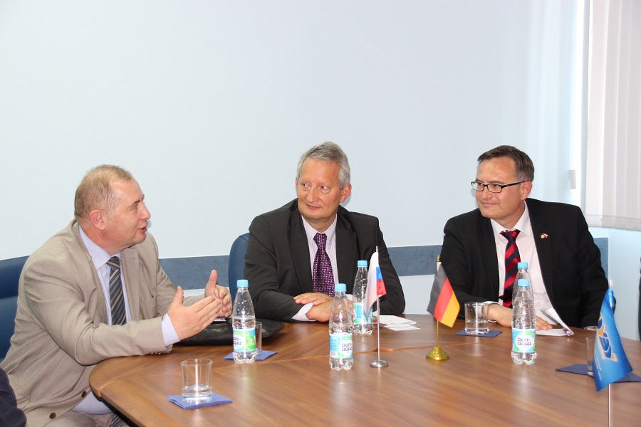 Drei Männer in Anzügen sitzen an einem mit kleinen internationalen Flaggen und Wasserflaschen geschmückten Konferenztisch und unterhalten sich in einem Büro an der Hochschule Coburg.