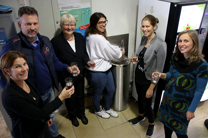 Eine Gruppe von sechs Personen steht in einem Raum der Hochschule Coburg, hält Gläser mit Wasser in den Händen und lächelt. Sie haben sich um einen Wasserspender versammelt, im Hintergrund sind Plakate und ein Getränkeautomat zu sehen.