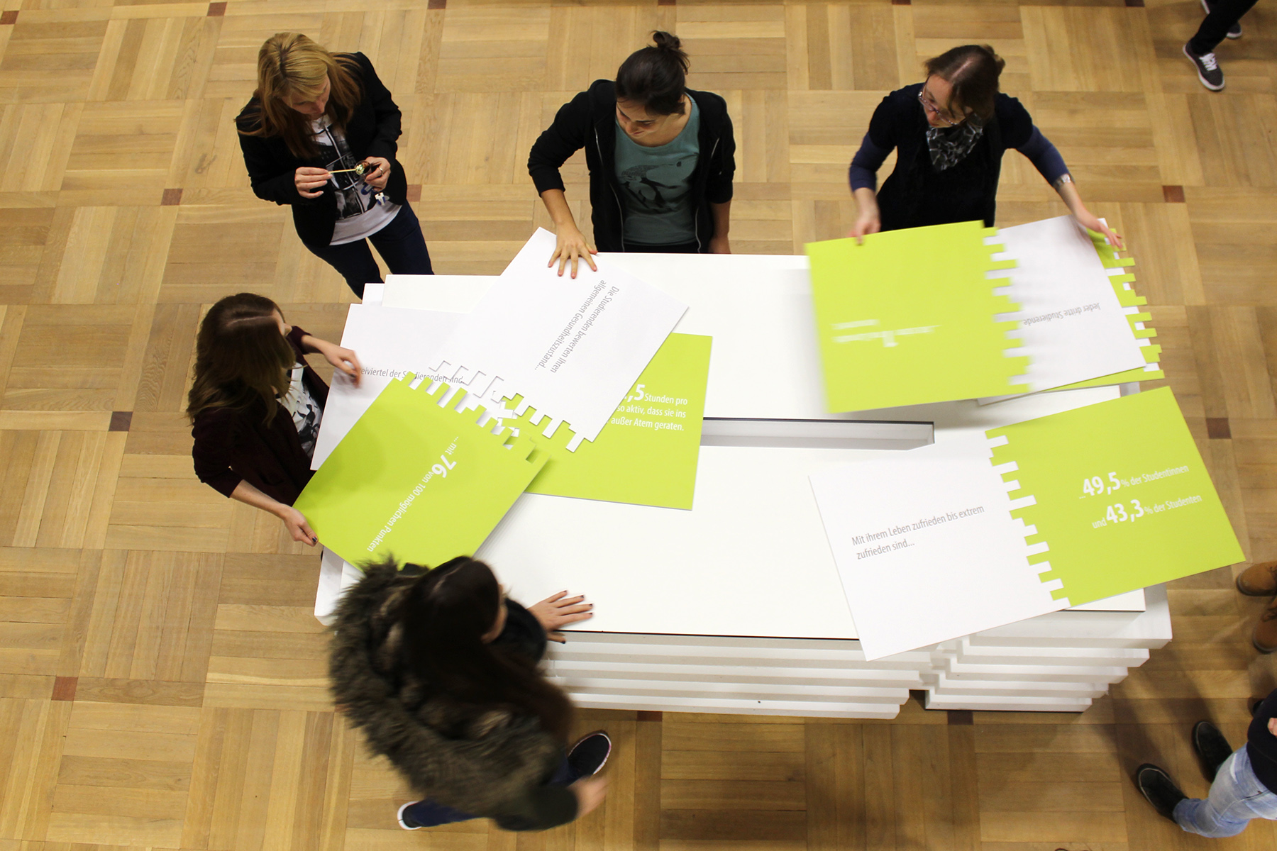 Eine Gruppe von Menschen, die den Geist der Hochschule Coburg verkörpern, steht um einen Tisch und ordnet große Puzzleteile. Die grünen rechteckigen Teile mit weißem Text liegen auf einem Holzboden und fördern eine Atmosphäre, die sowohl kollaborativ als auch konzentriert ist.