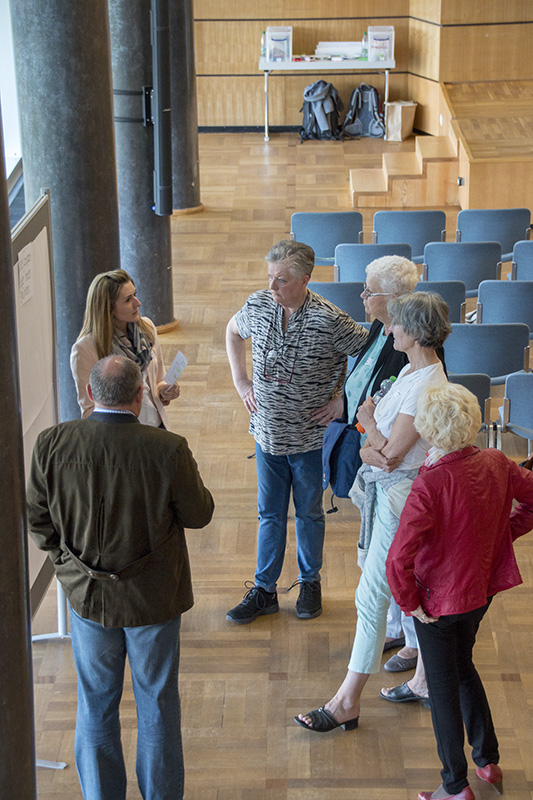 Eine Gruppe von Menschen steht in der geräumigen Halle der Hochschule Coburg und unterhält sich neben einer großen Tafel. Die Kulisse mit Reihen leerer Stühle und Holzboden schafft eine formelle und doch lockere Atmosphäre.